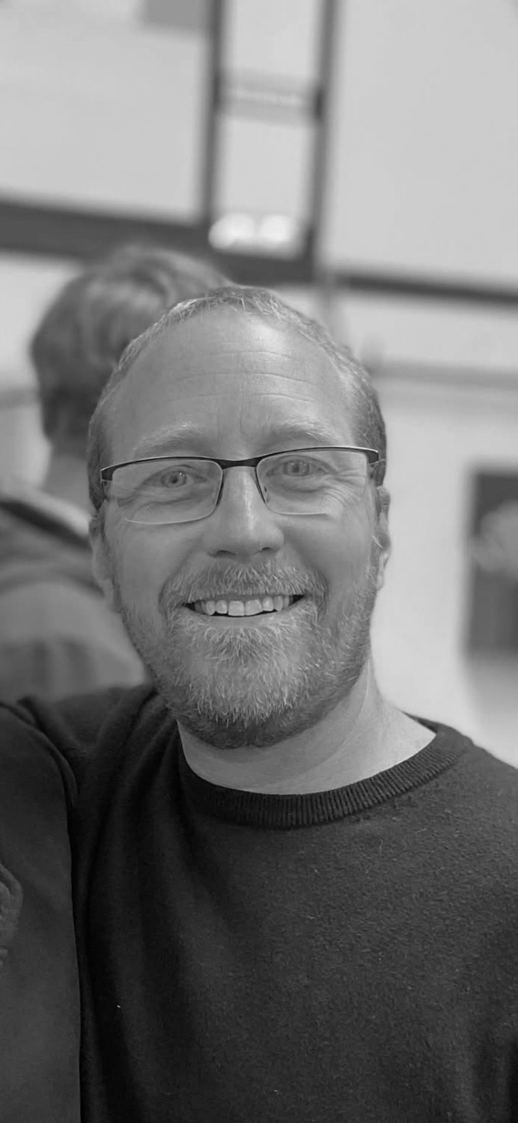 Black and white photo of a smiling man with glasses and a beard, wearing a dark sweater, in an indoor setting with a blurry background.