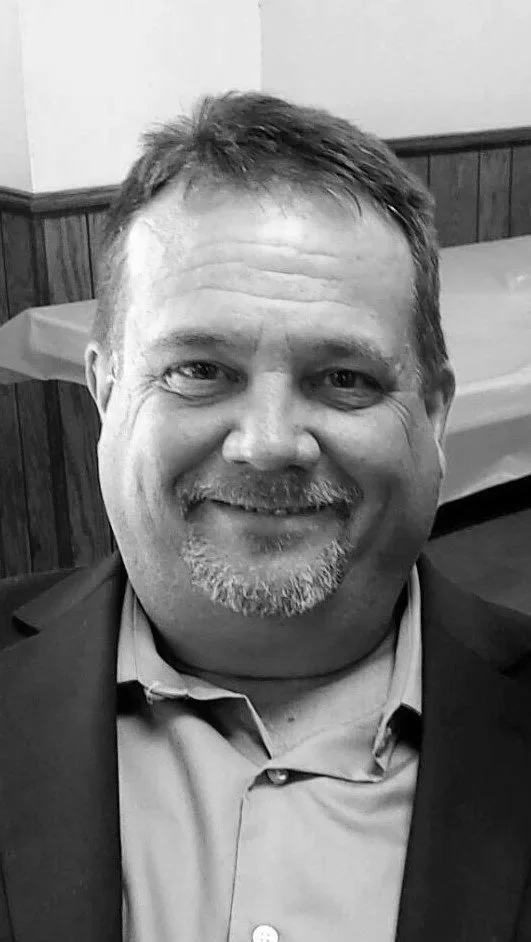 A smiling man with short hair, a beard, and a mustache, wearing a suit jacket and a button-up shirt, indoors with wooden paneling and a table in the background.