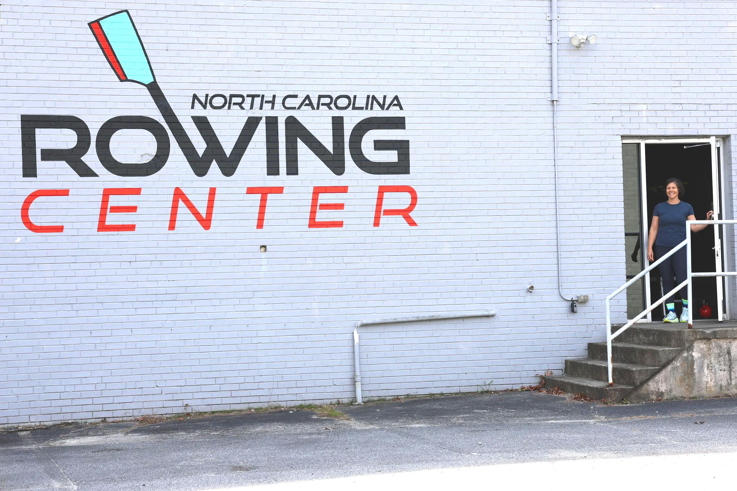A woman standing at the entrance of the North Carolina Rowing Center, smiling, with a mural of a boat paddle and the words "North Carolina Rowing Center" on the brick wall behind her.