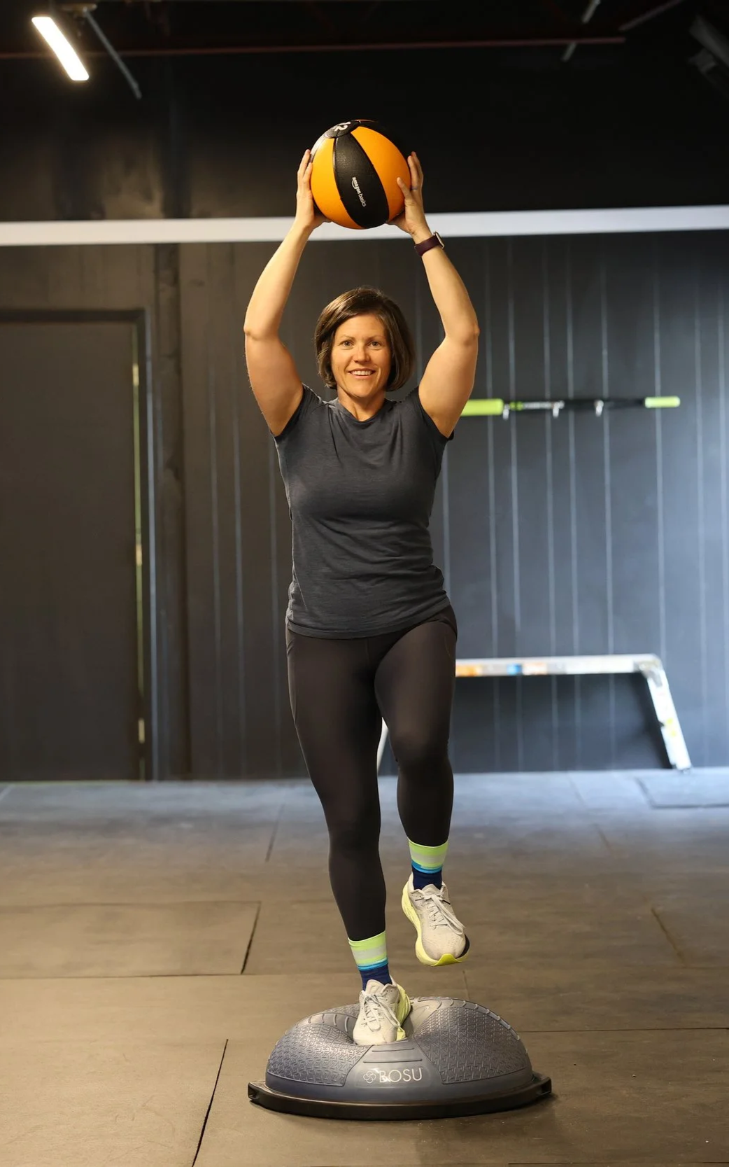 A woman in workout clothes is standing on a BOSU ball while holding a medicine ball above her head in a gym.