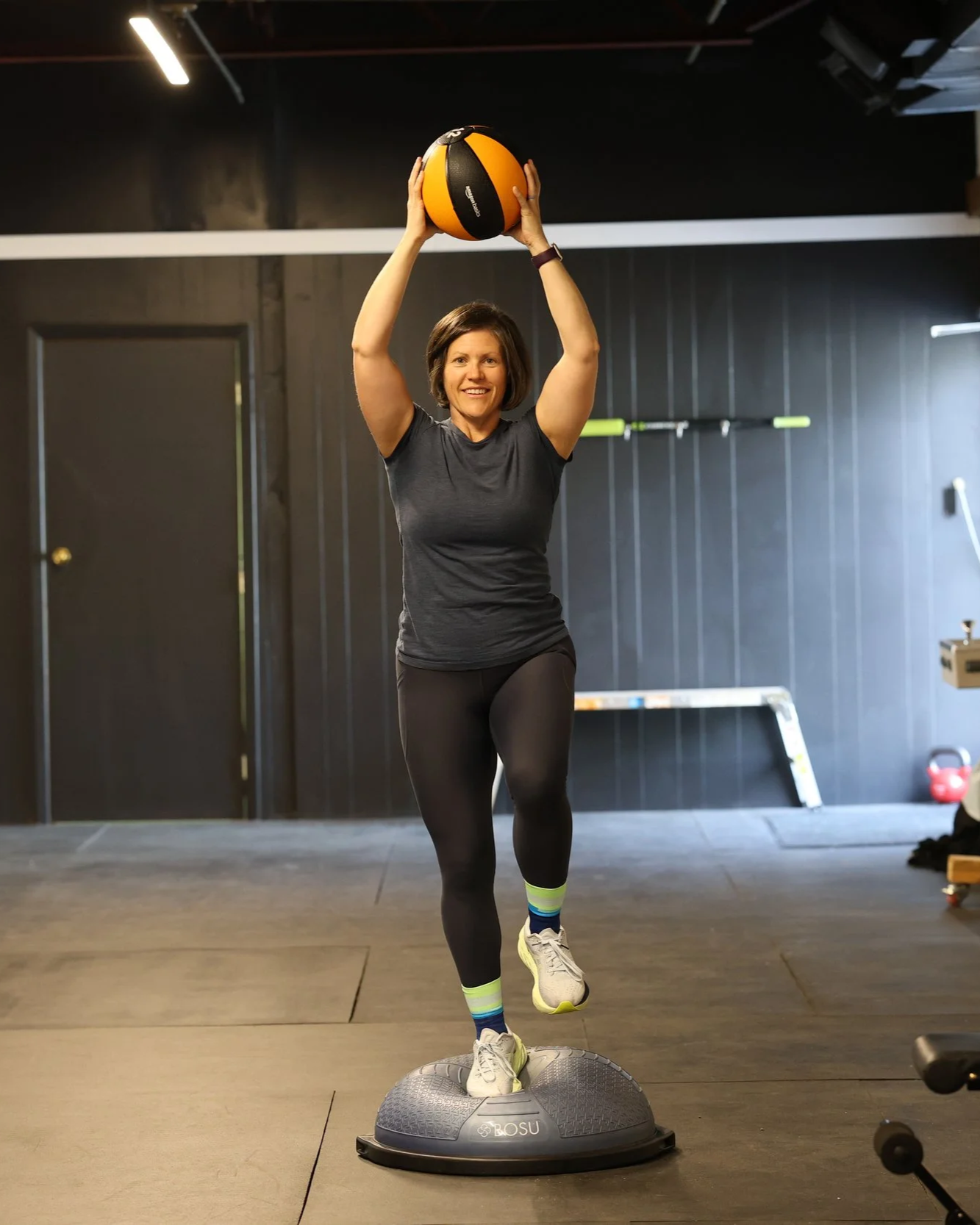A woman in workout clothes is balancing on one foot on a BOSU ball while holding a medicine ball above her head in a gym.