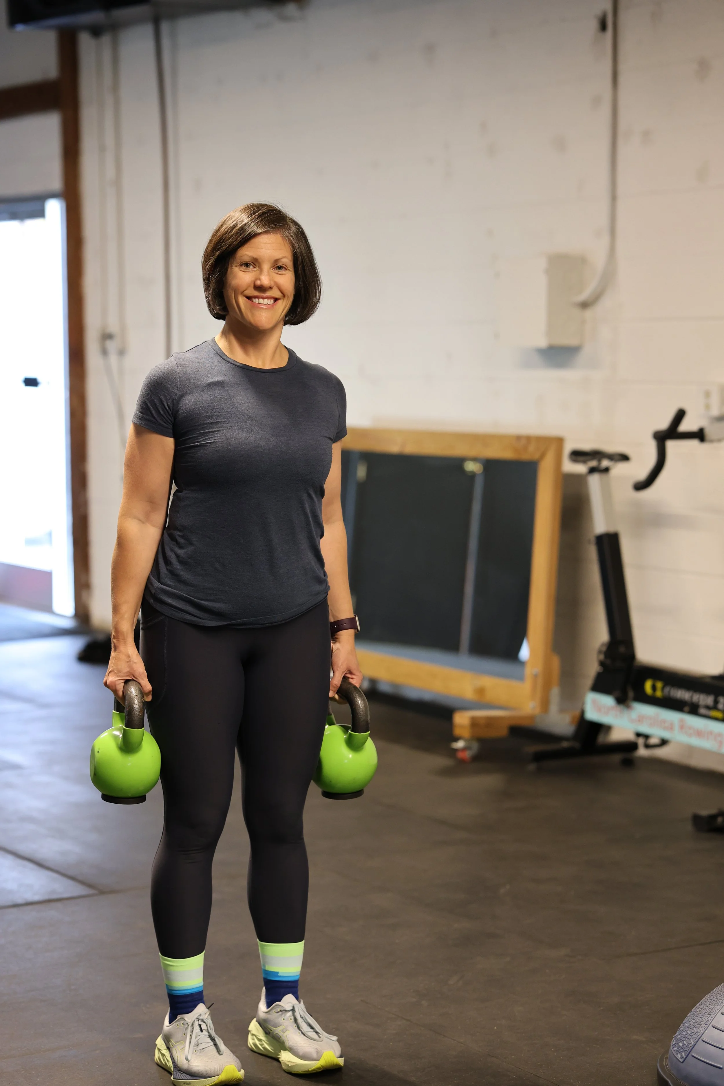 A woman holding two green kettlebells in an indoor gym.