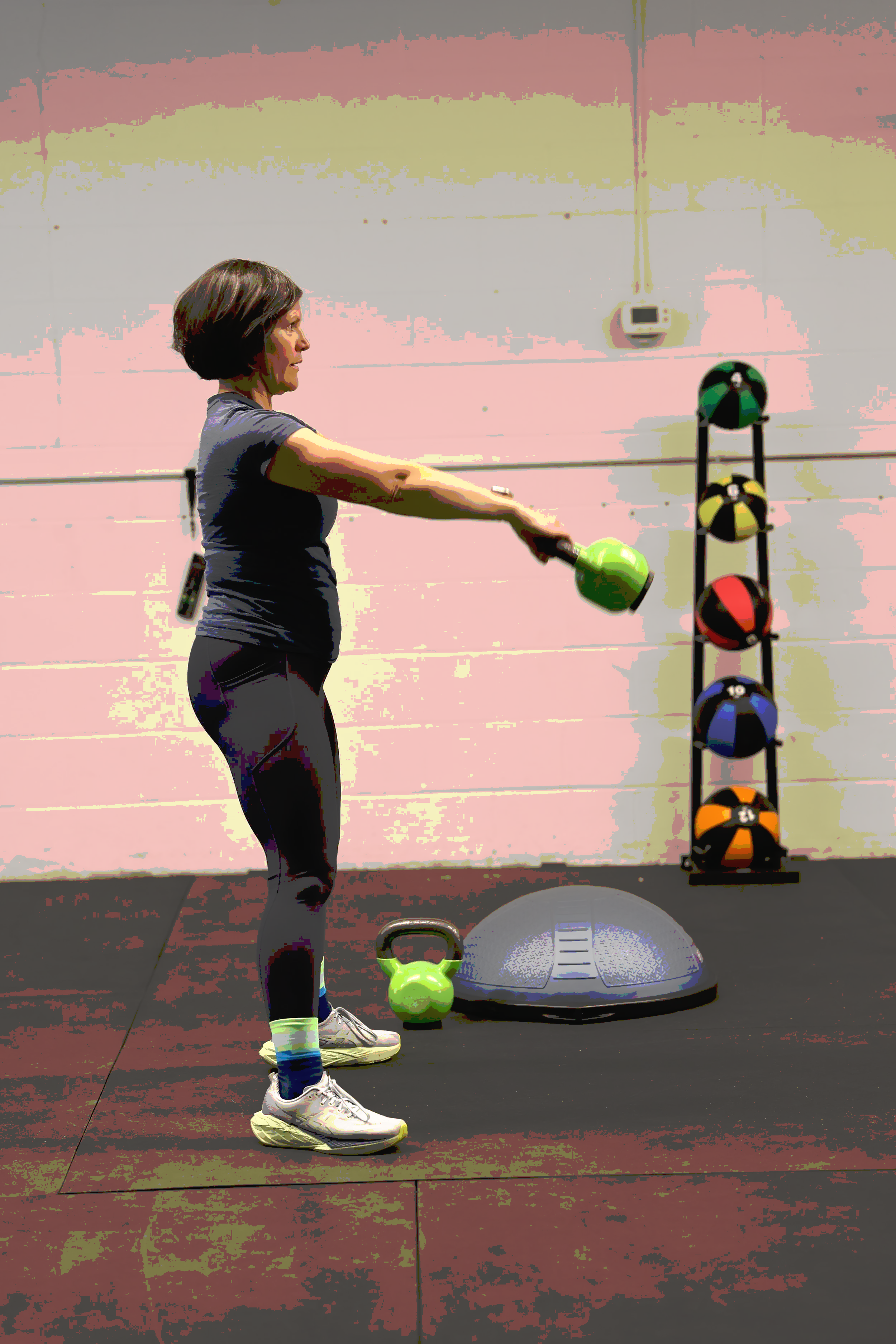 Caroline Duncan lifting a green kettlebell in a gym with fitness equipment including a balance trainer and a rack of medicine balls in the background.