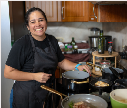 A woman smiling and cooking in a kitchen, wearing a black apron.