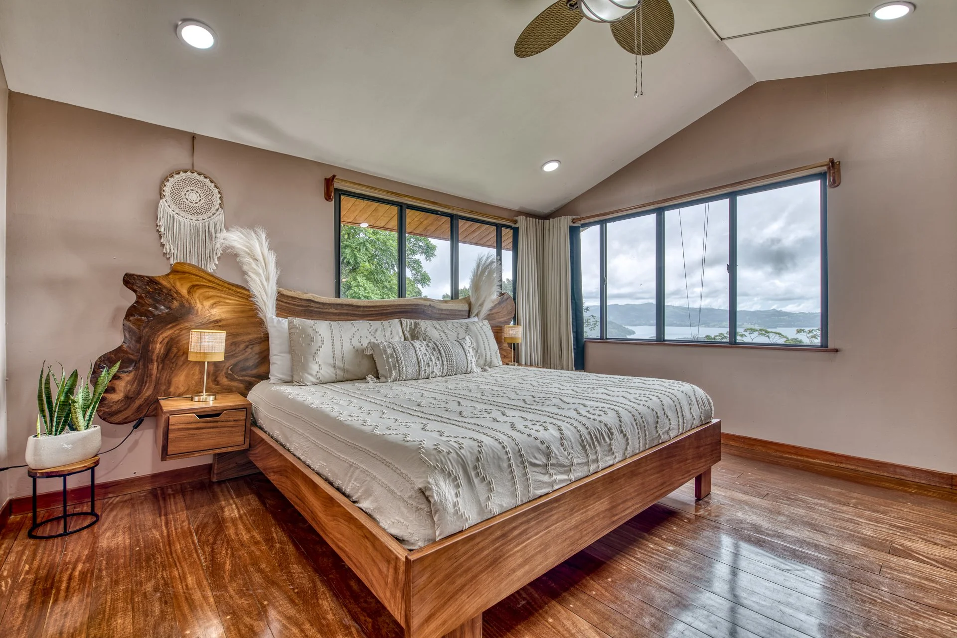 Bedroom with a large wooden bed, decorative pillows, small bedside lamp, potted plant, beige walls, large windows showing a lake and mountains, ceiling fan, wooden flooring.