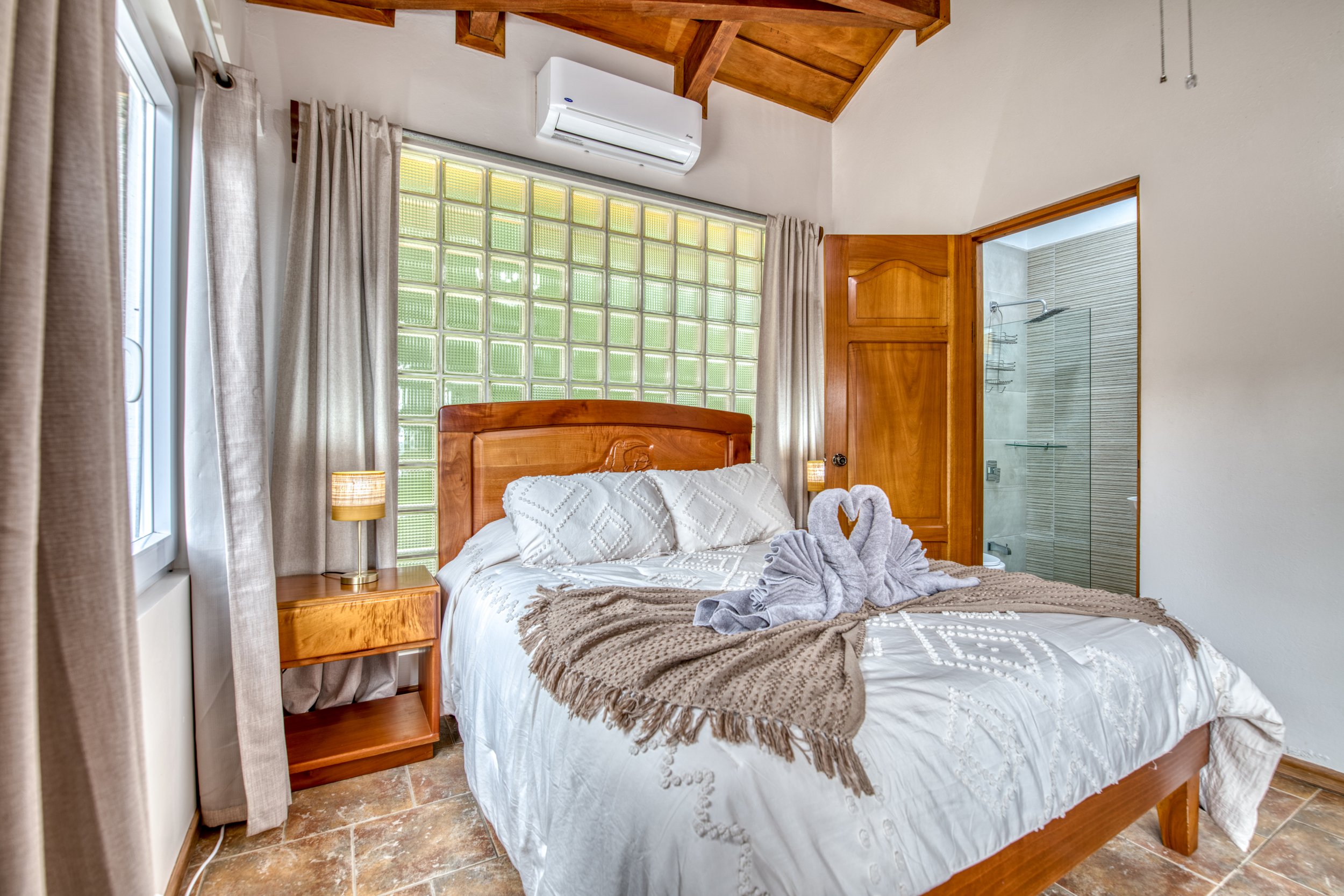 A cozy bedroom with a wooden bed, white bedding, and towel swans on the bed. There is a window with beige curtains, a nightstand with a lamp, a window with glass blocks behind the bed, and an open door to a bathroom with tiled walls and shower.