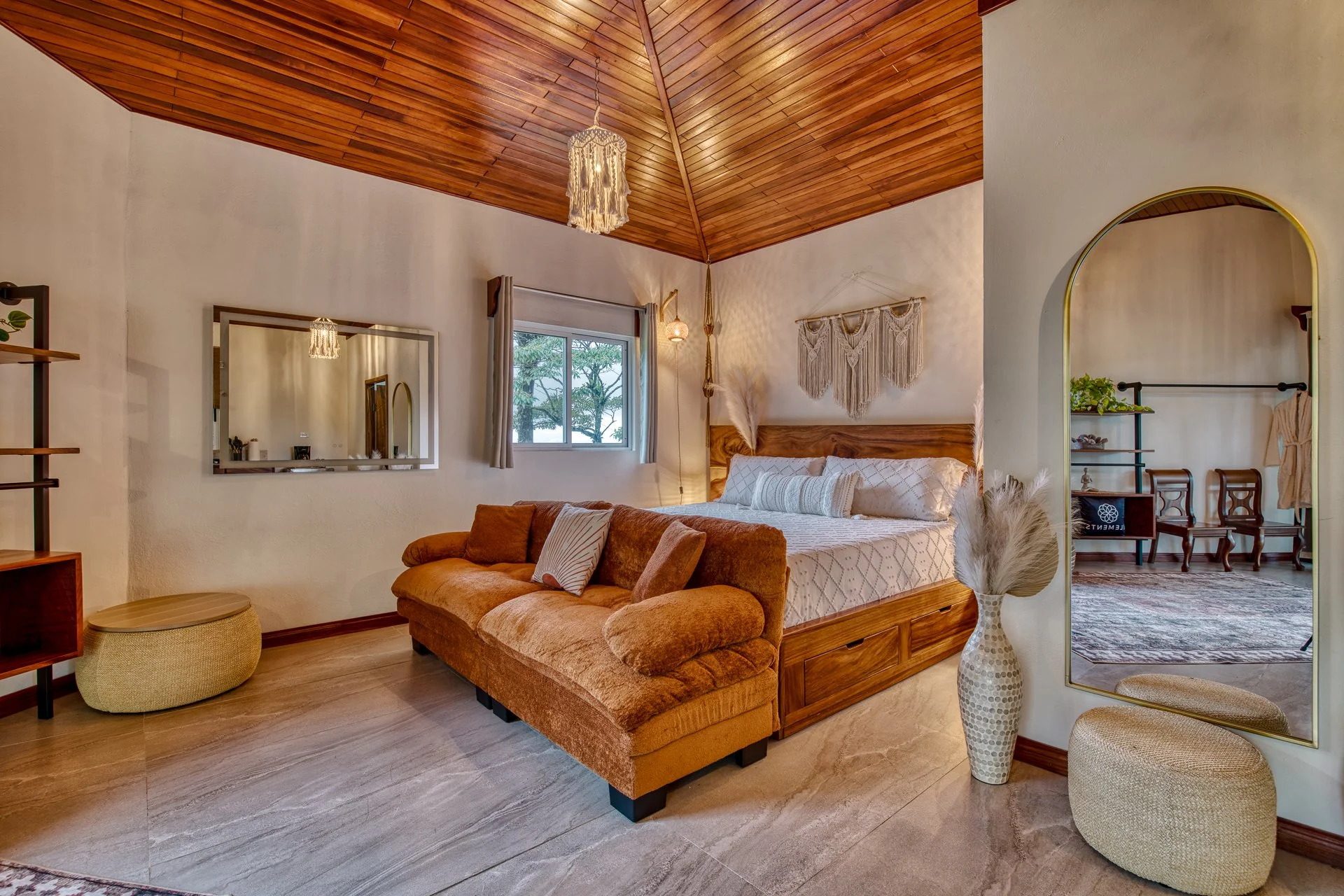 A cozy bedroom with a wooden ceiling, a large bed with white bedding, a beige armrest sofa at the foot of the bed, a window with curtains, a wall mirror, a hanging lamp, wall art, a vase with pampas grass, and an open doorway leading to another room with chairs and a rug.