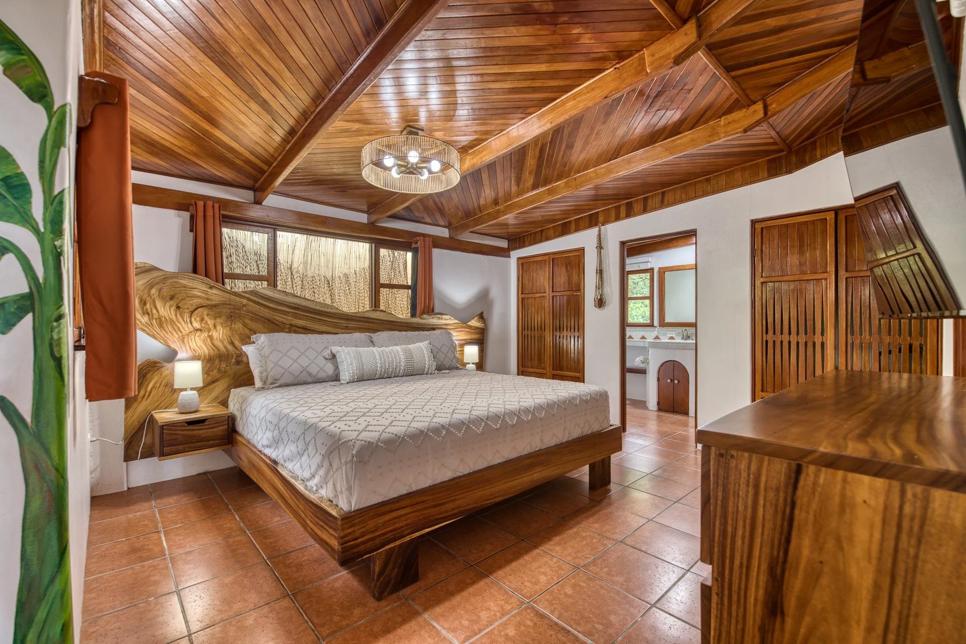 Interior of a bedroom with a wooden ceiling, large bed with patterned bedding, small bedside lamps, and wooden furniture, with the bathroom visible through an open door.