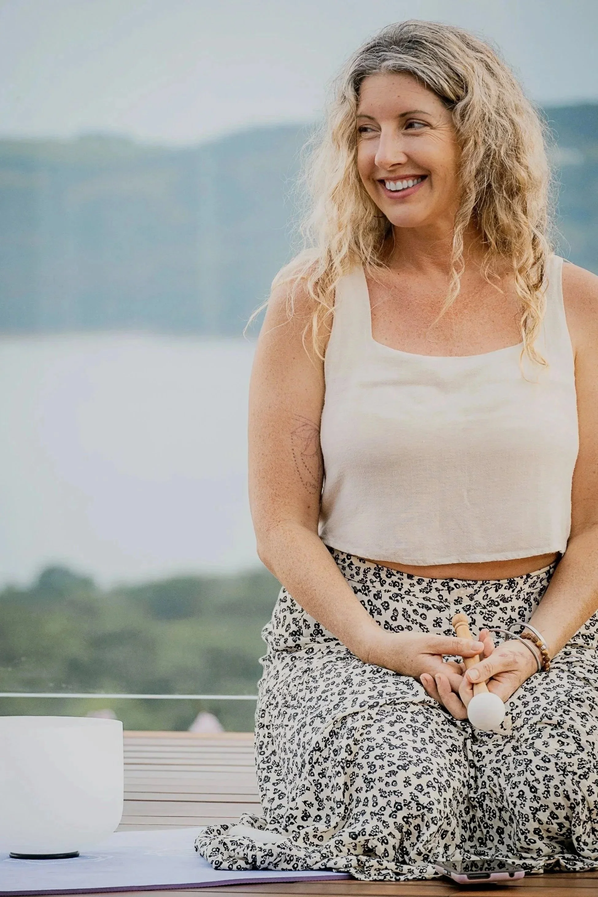 A woman with curly blonde hair smiling while sitting outdoors, with mountains and a lake in the background.