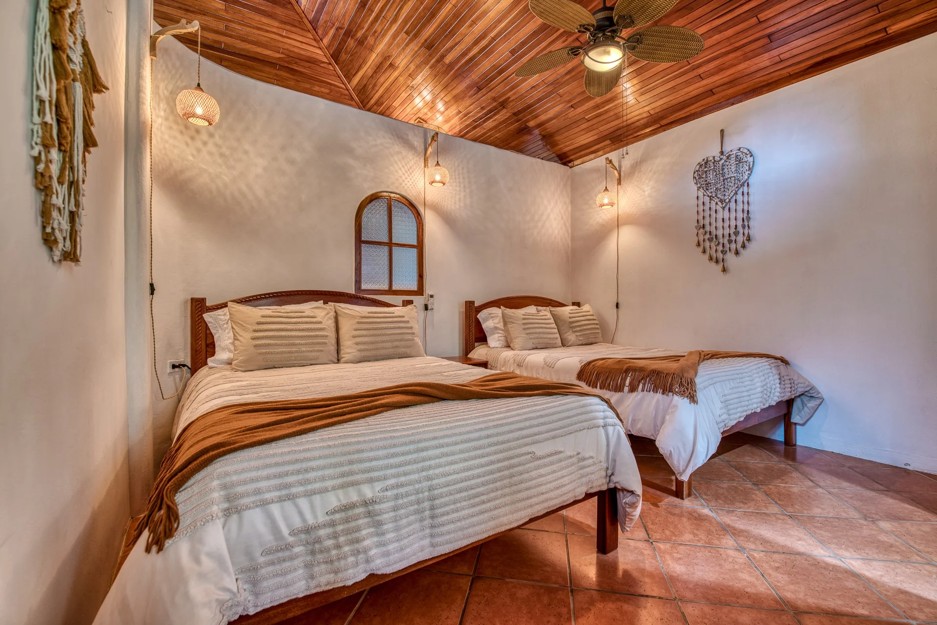 A bedroom with wooden ceiling and tile floor, two beds with beige bedding, decorative pillows, and brown throws, hanging wall decorations, a ceiling fan, and soft lighting.