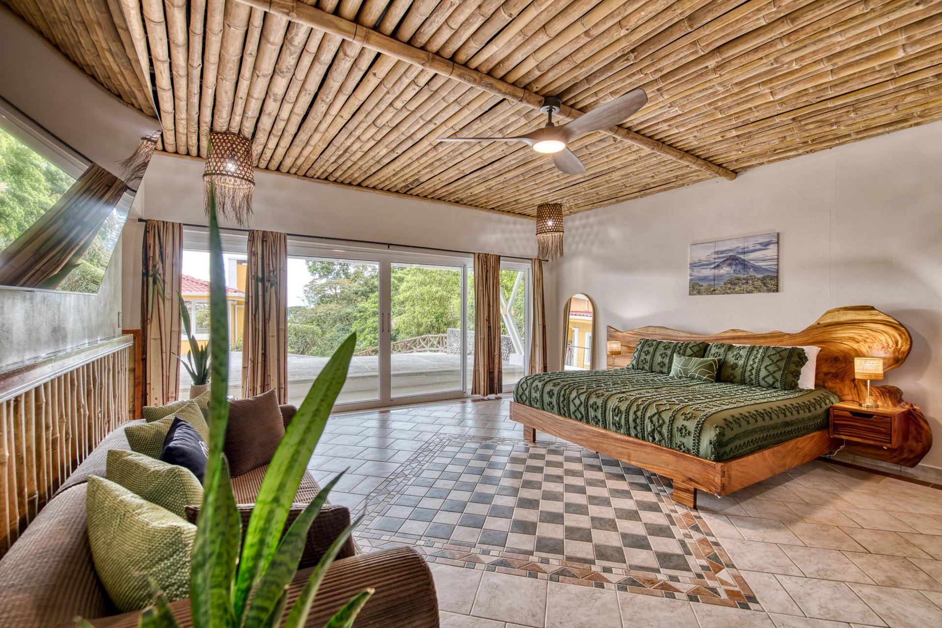 A tropical hotel room with a large wooden bed, patterned green bedding, a natural wood headboard with curved edges, side tables with lamps, a sliding glass door leading to a balcony, ceiling fan, bamboo ceiling, and curtains with a nature view outside.