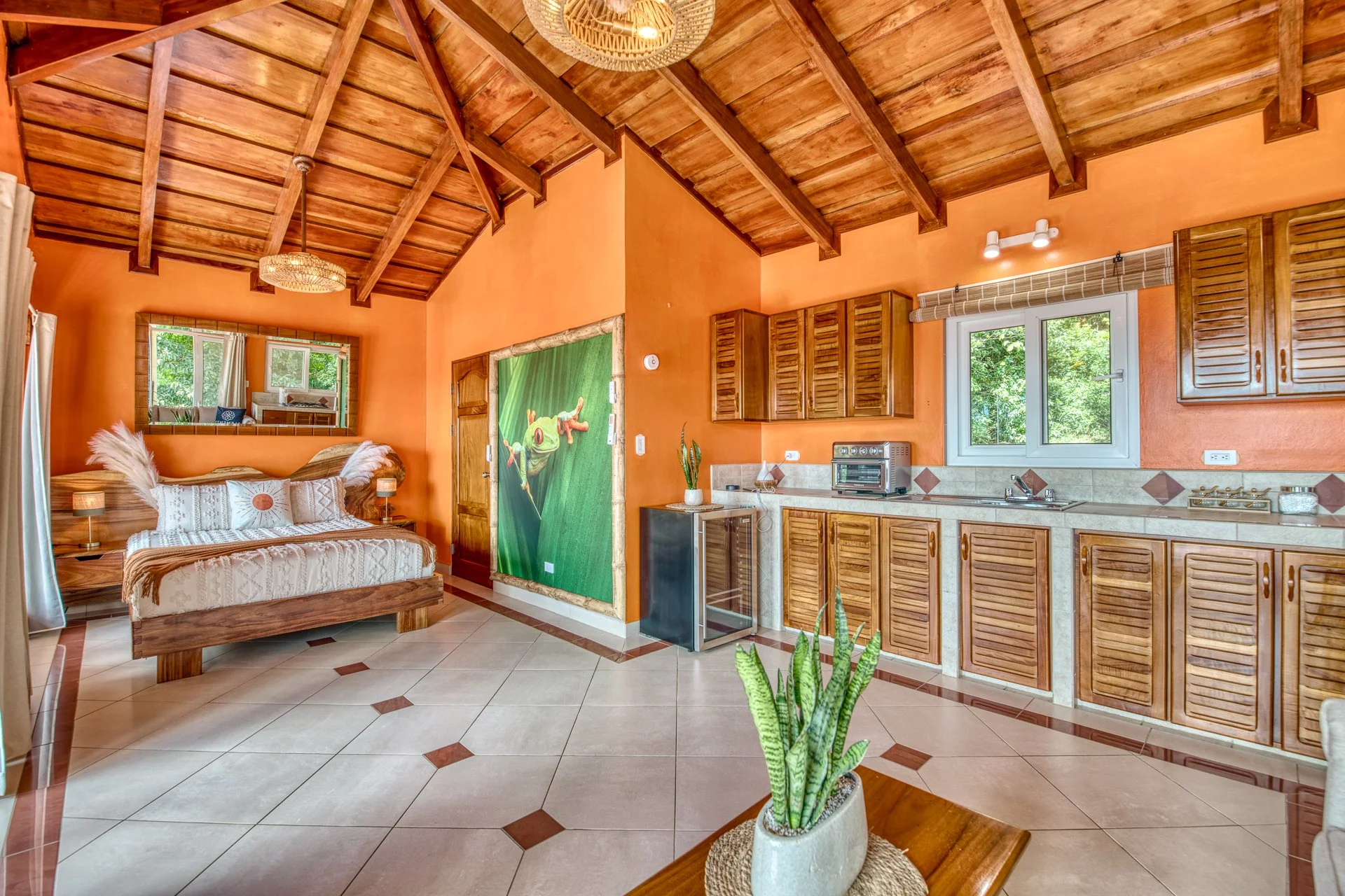 A cozy room with a wooden vaulted ceiling, orange walls, and a large mirror above a bed with white bedding and feather pillows. The room features tiled floors, a kitchenette with wooden cabinets, a small window showing greenery outside, and a small black mini-fridge. A potted snake plant is on a wooden table in the foreground.