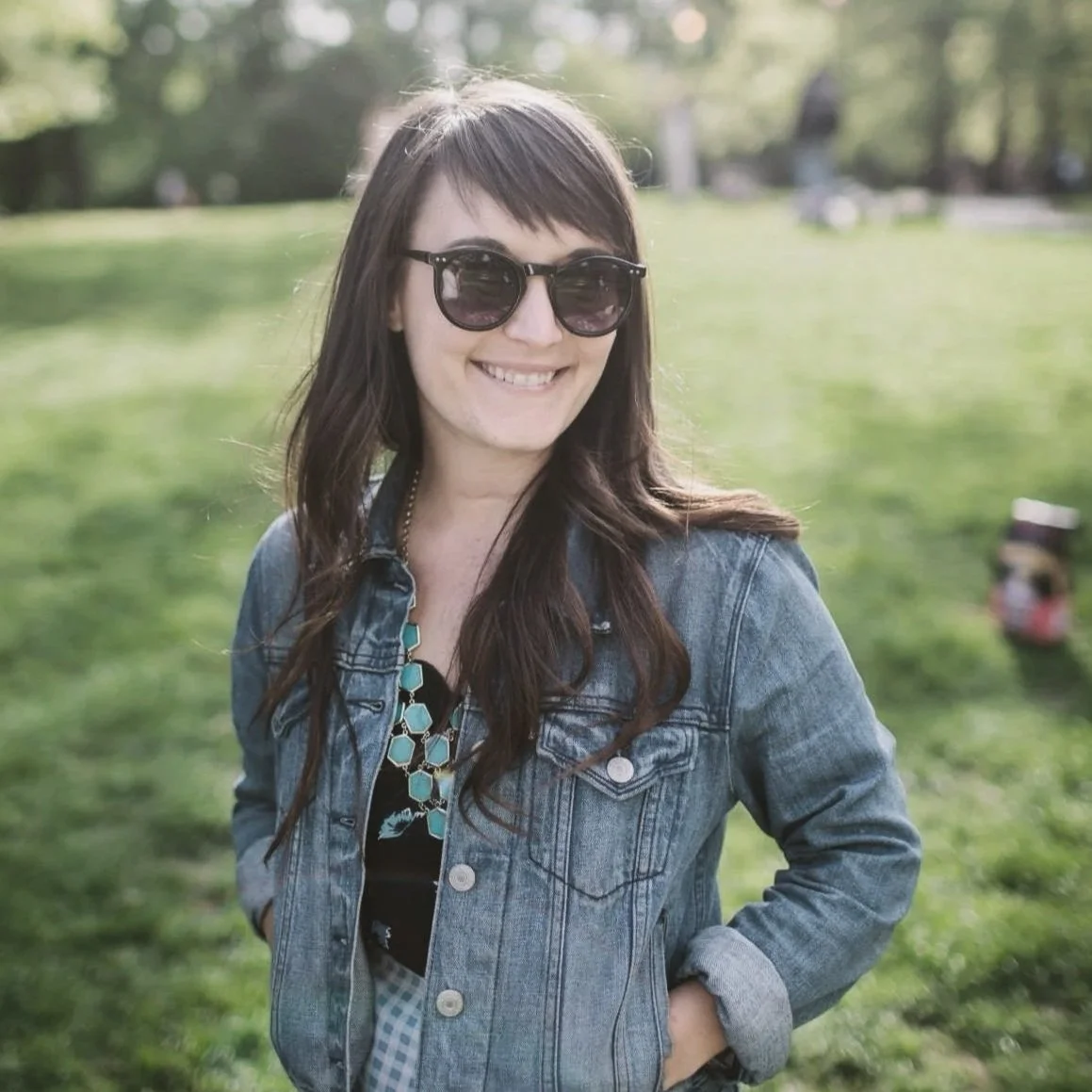 A woman with long dark hair wearing sunglasses, a denim jacket, and a turquoise necklace, smiling and standing outdoors in a park with green grass and trees in the background.
