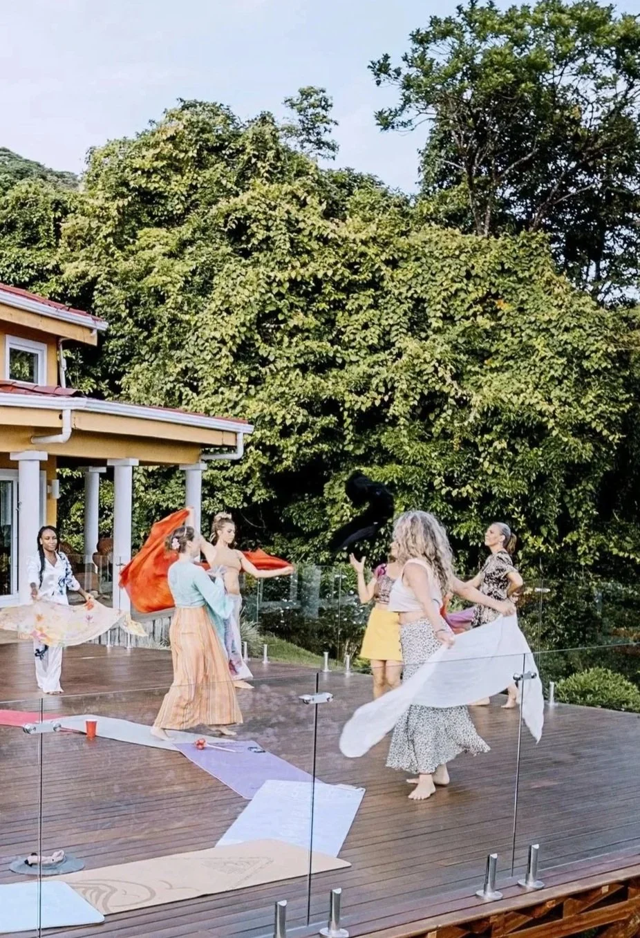 Women dancing on a wooden deck outdoors, surrounded by green trees, with some holding or twirling colorful fabrics.