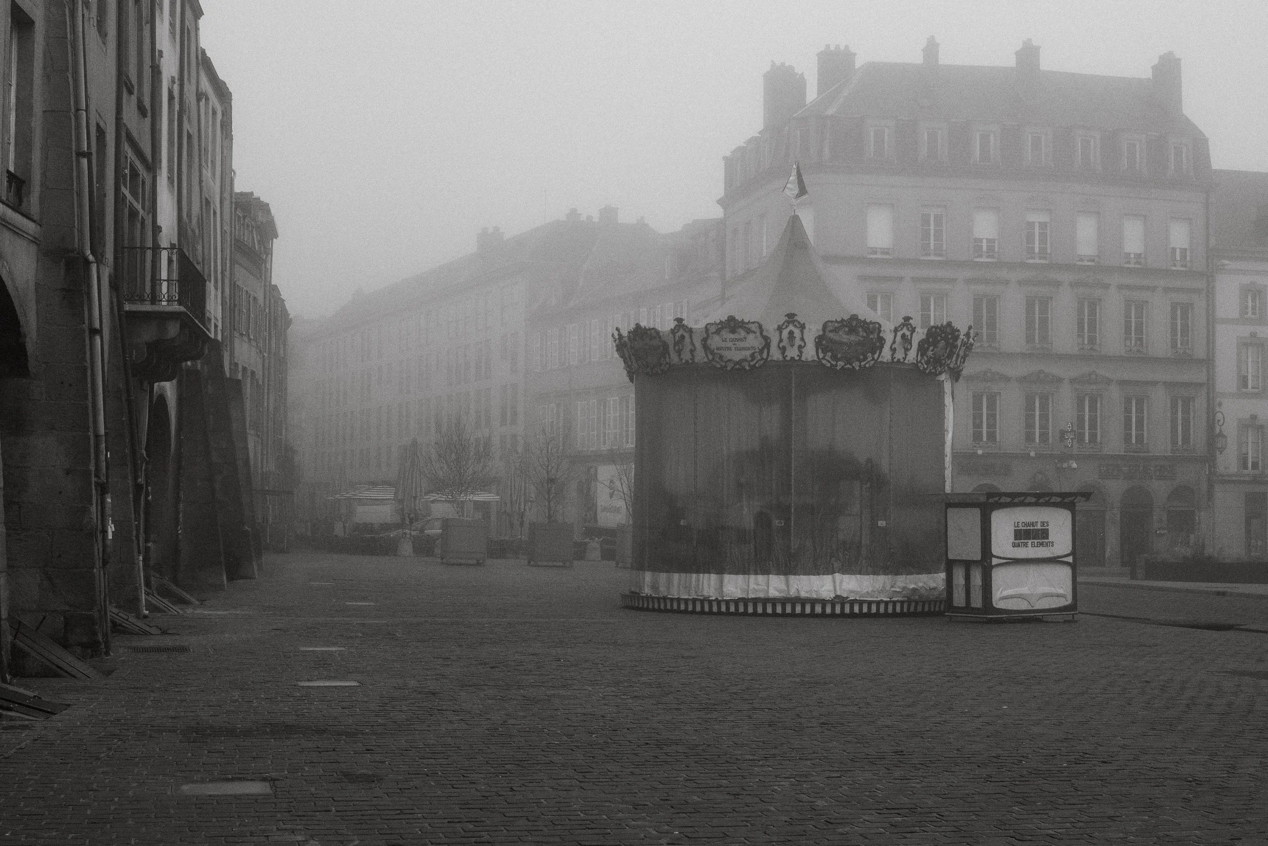 Une place pavée vide avec un manège de carnaval au centre, entouré de bâtiments anciens sous un brouillard.