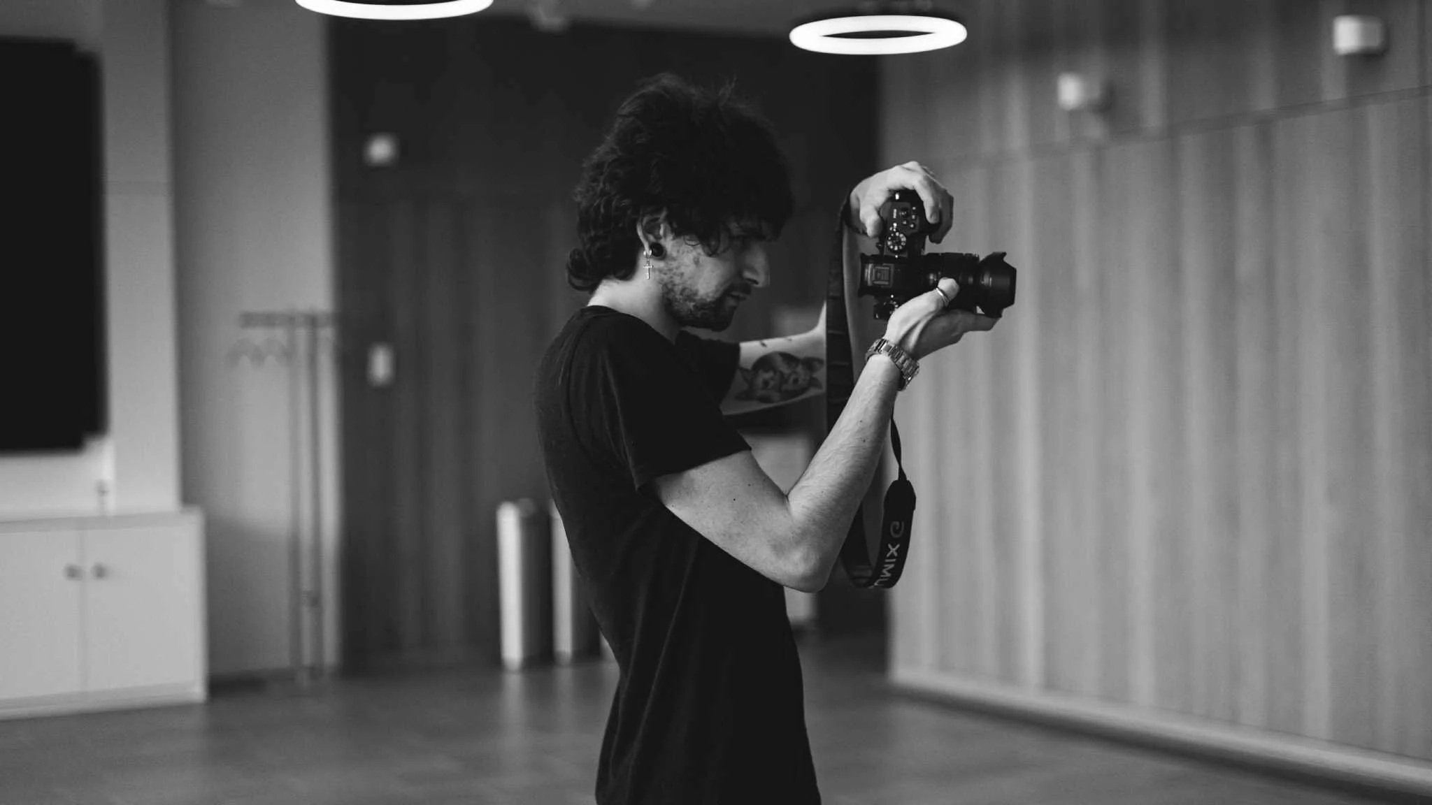 Un homme avec des cheveux bouclés, portant un t-shirt noir, prend une photo avec un appareil photo dans une pièce au mur en bois, vue en profil. L'atmosphère semble artistique ou créative.