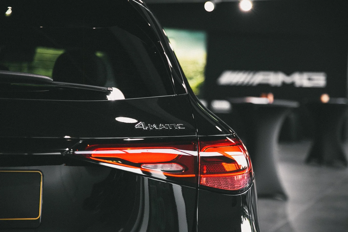 Vue de l'arrière d'une voiture noire Audi avec le badge '4 MATIC' et les feux arrière allumés, dans un garage ou un espace intérieur avec un grand logo Mercedes-AMG en arrière-plan.