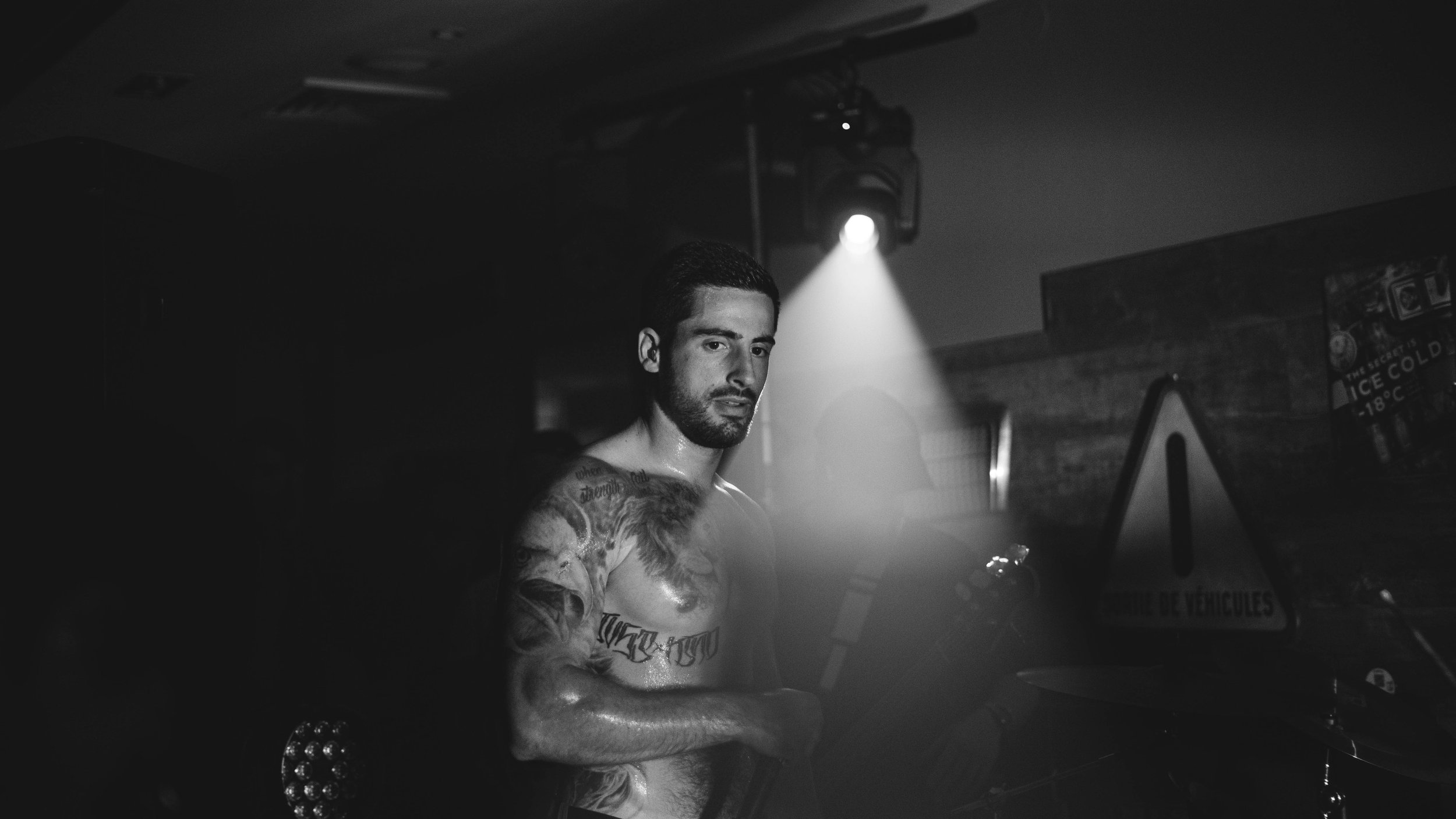 Un homme tatoué derrière un DJ dans une ambiance sombre et éclairée par un projecteur, en noir et blanc.
