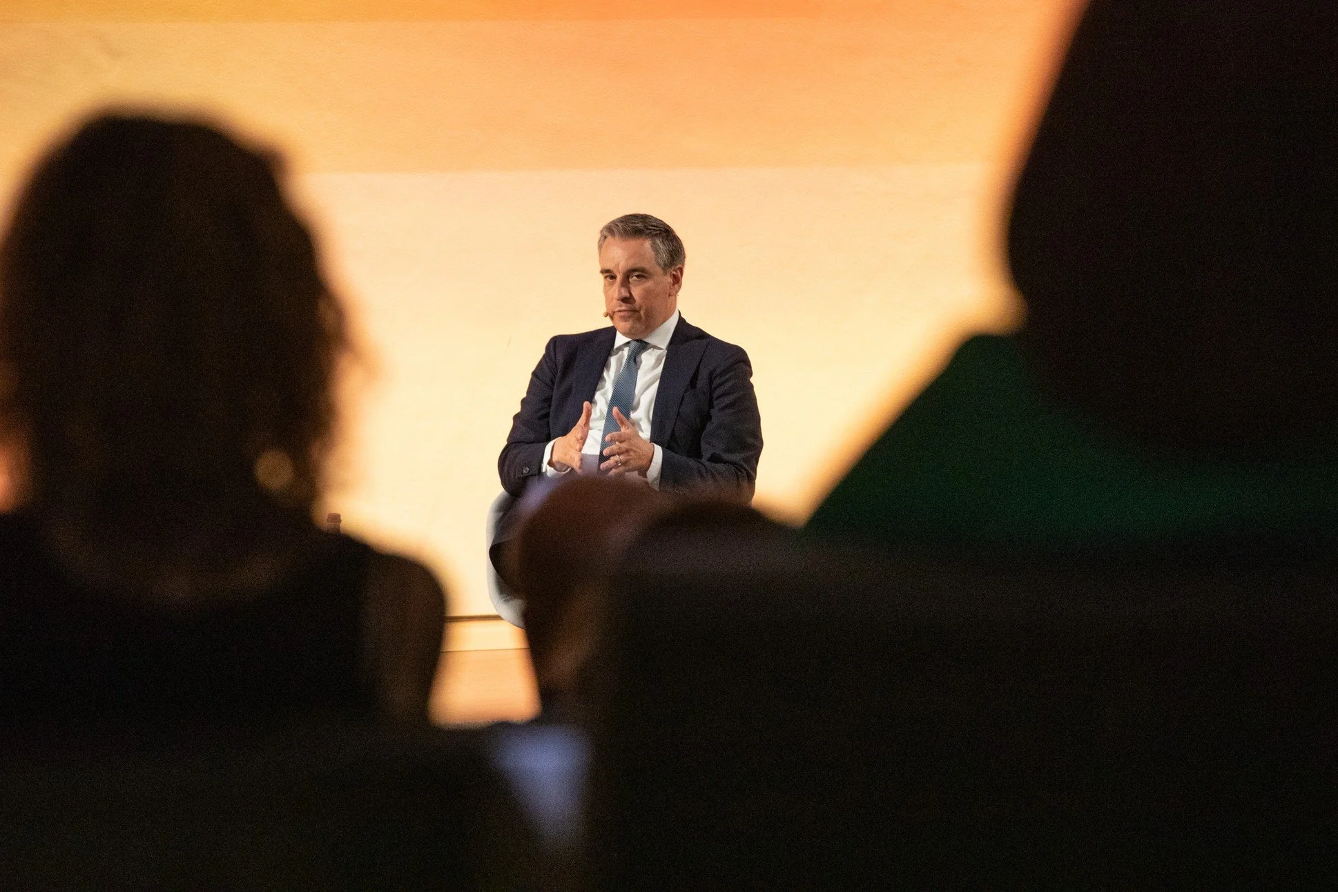 Homme en costume assis sur une scène, parlant lors d'une conférence, vue à travers les silhouettes du public.