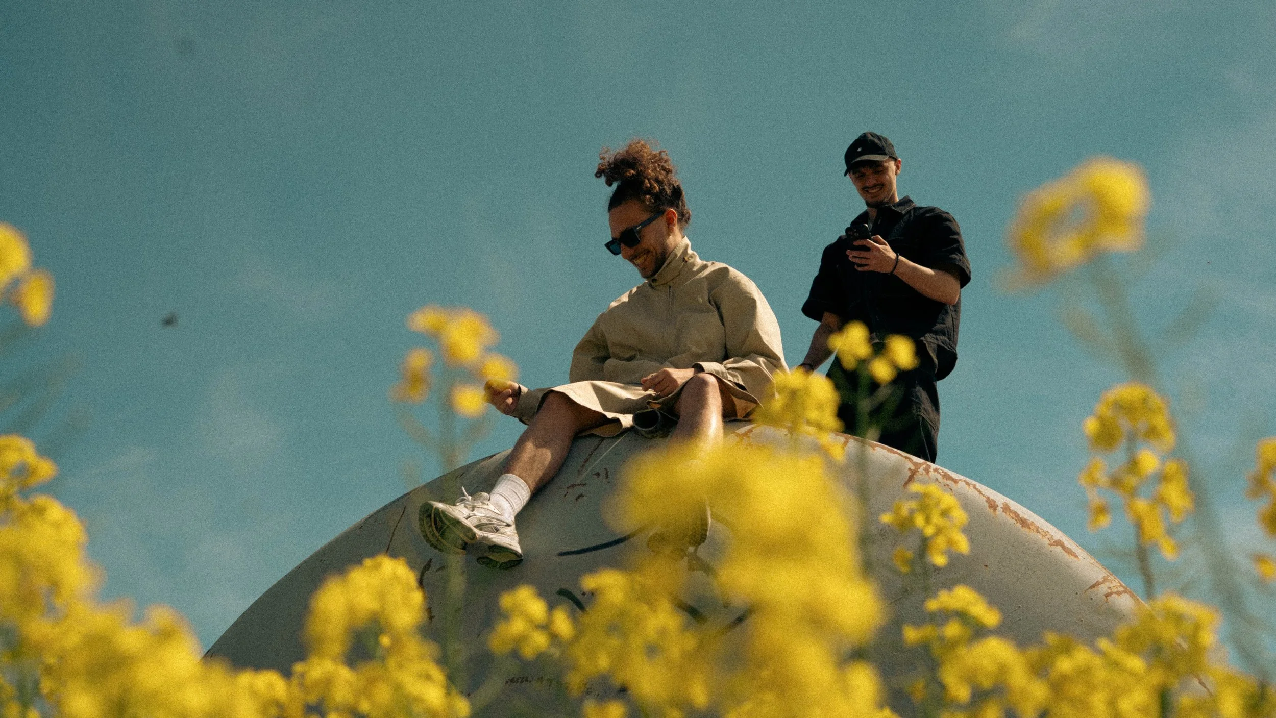 Deux jeunes hommes assis sur une structure métallique gonflée, entourés de fleurs jaunes, avec un ciel clair en arrière-plan. L’un d’eux regarde son téléphone, l’autre regarde le sien, tous deux souriants.