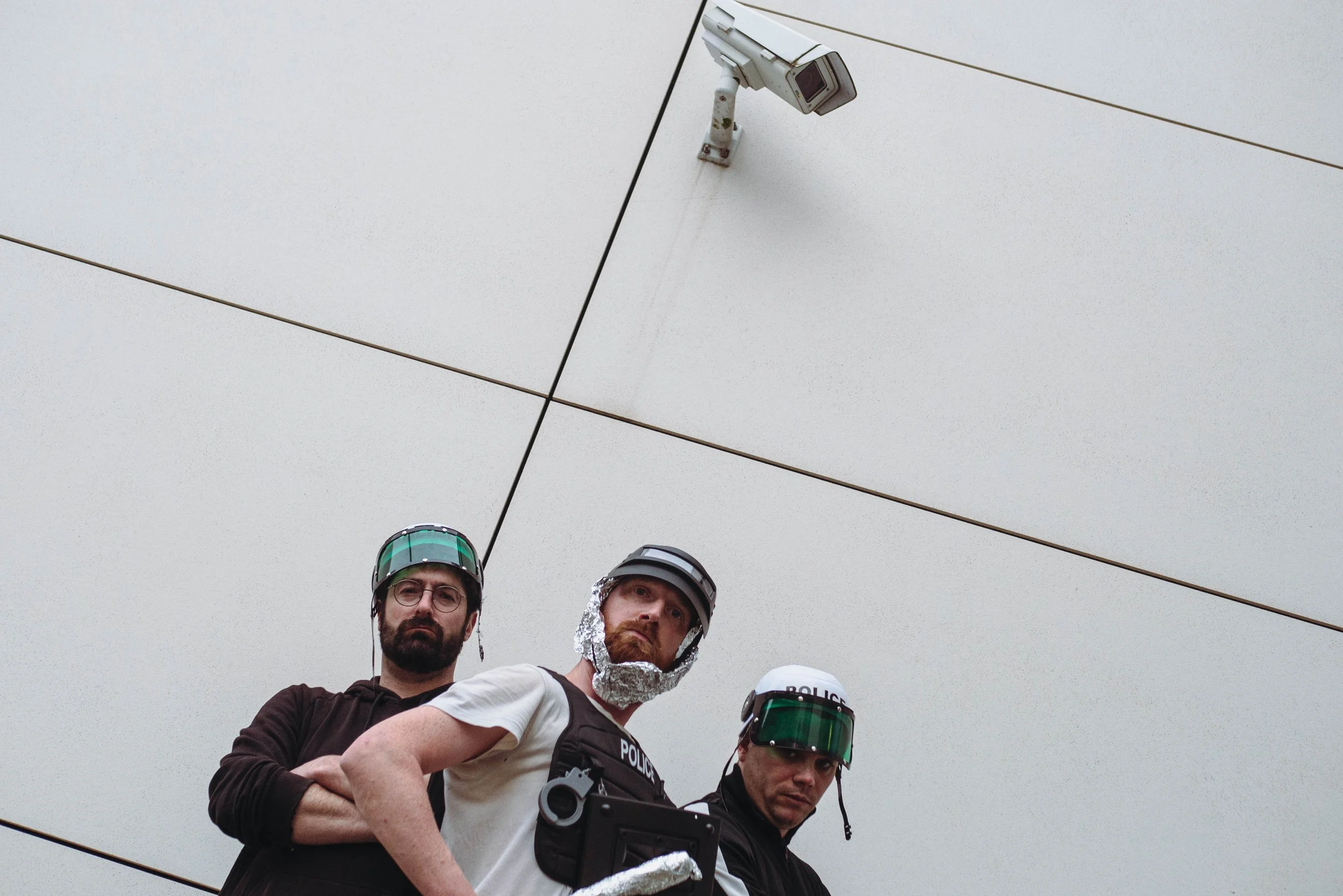 Trois hommes, dont deux policiers, portant des casques, au-dessus, une caméra de surveillance, le ciel est gris.