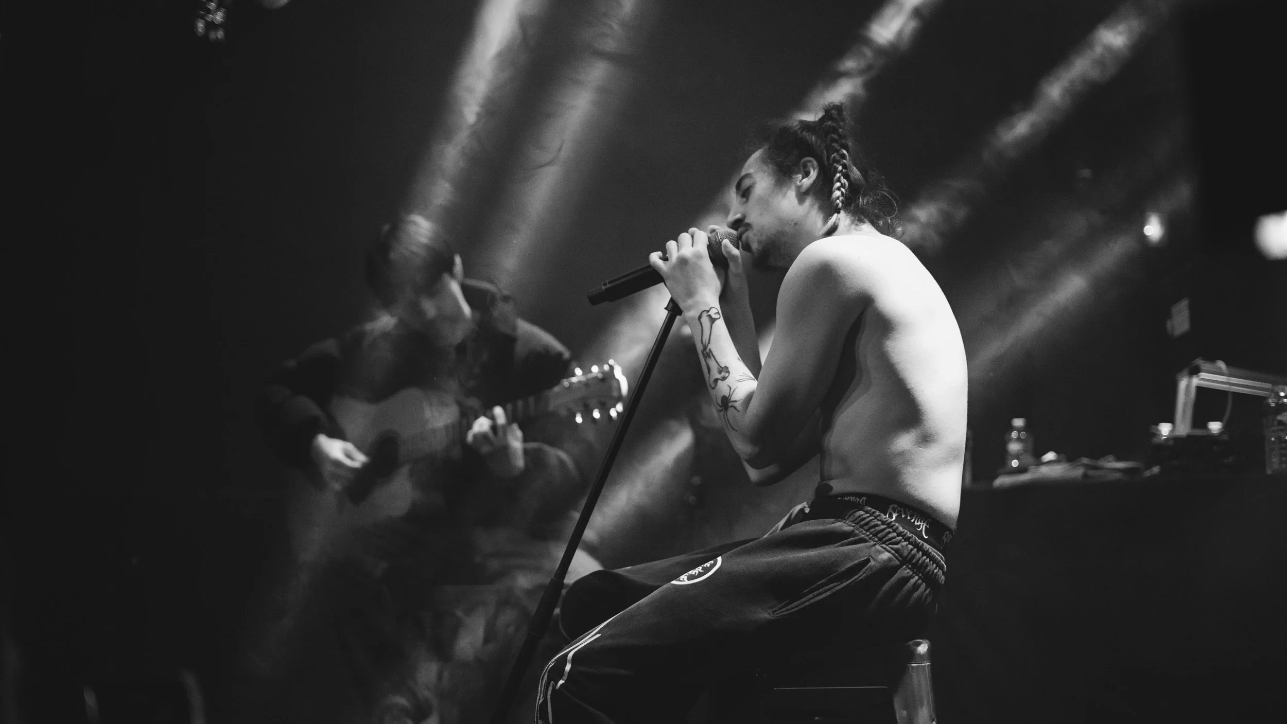 Un homme nu, avec des tatouages, chante en studio avec un microphone, tandis qu'une personne joue de la guitare en arrière-plan, dans une ambiance sombre avec des lumières.