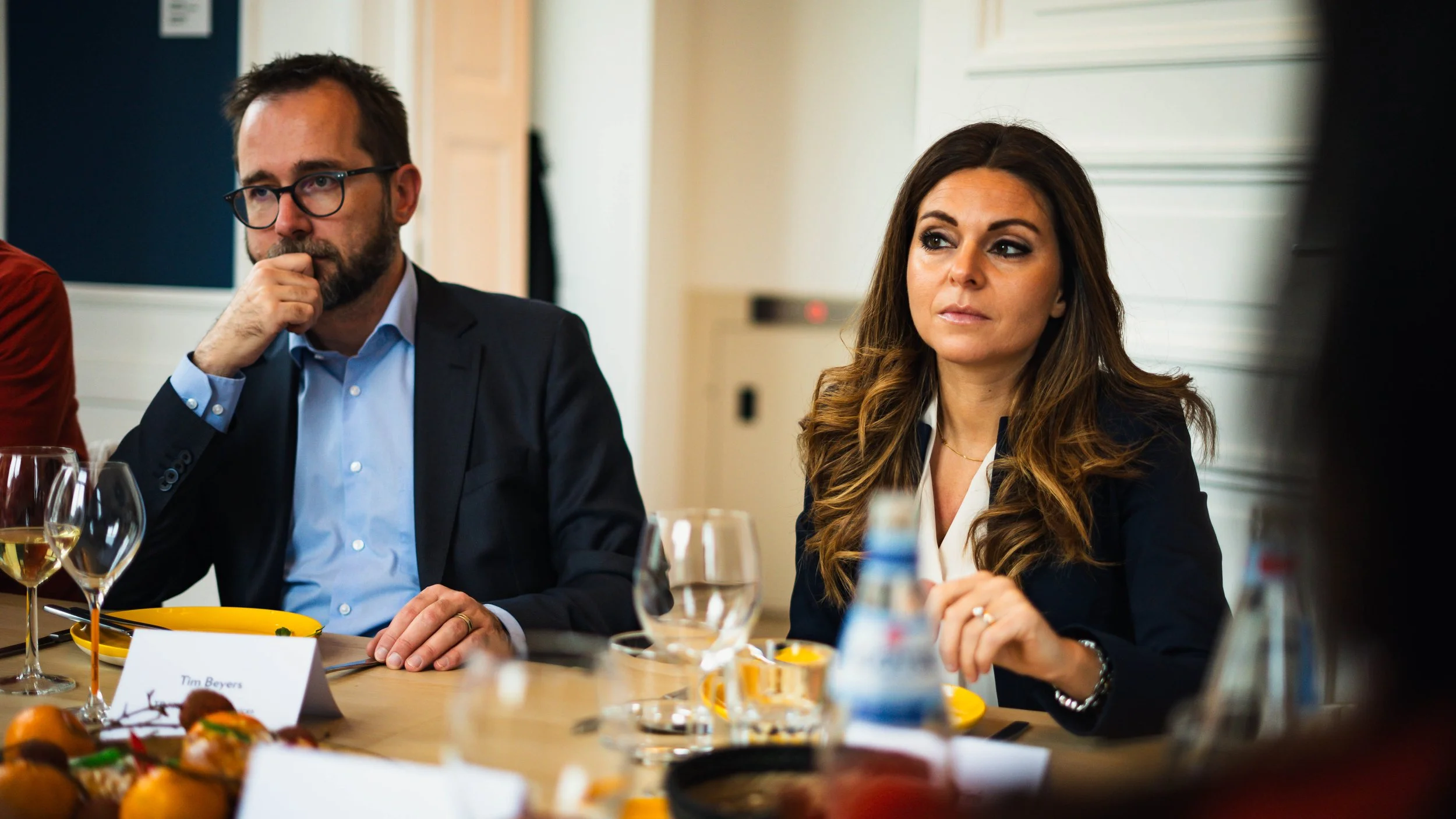Deux personnes assises à une table lors d'une réunion ou d'un repas formel, avec des verres de vin et des plats devant elles, dans une salle bien éclairée.