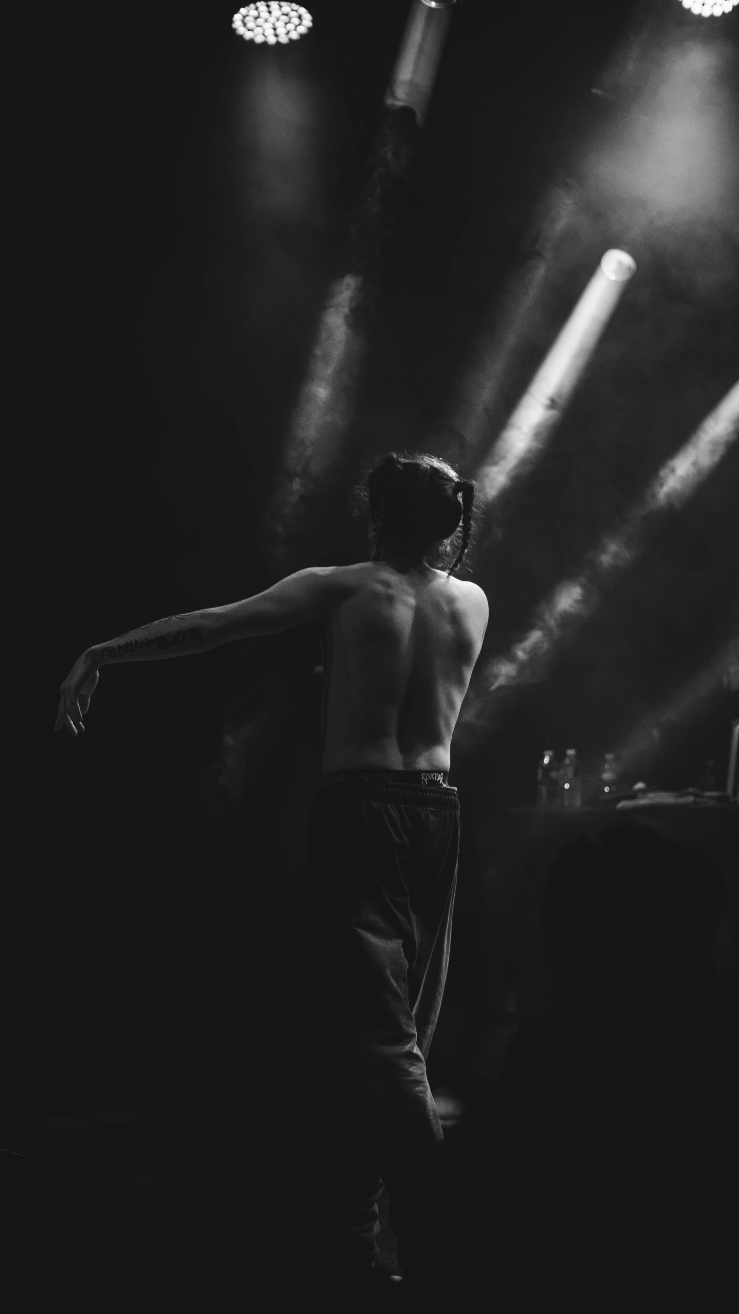 Une personne nue de dos, avec des cheveux tressés, danse dans une lumière sombre, sur scène avec des projecteurs à l'arrière-plan.