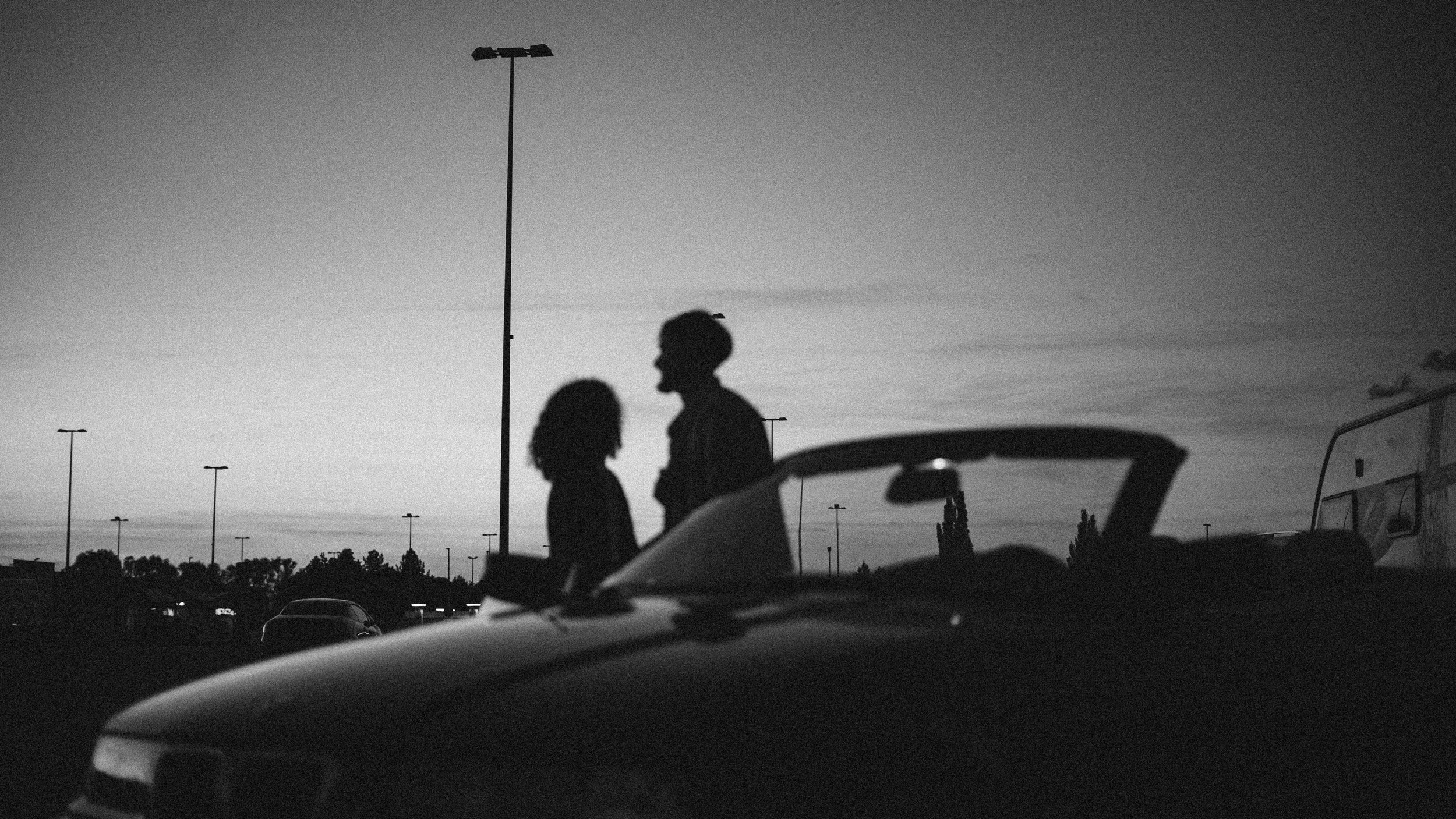 Silhouette de deux personnes échangeant dans un parking au coucher du soleil, avec une voiture rétro en premier plan et des lampadaires alignés en arrière-plan.