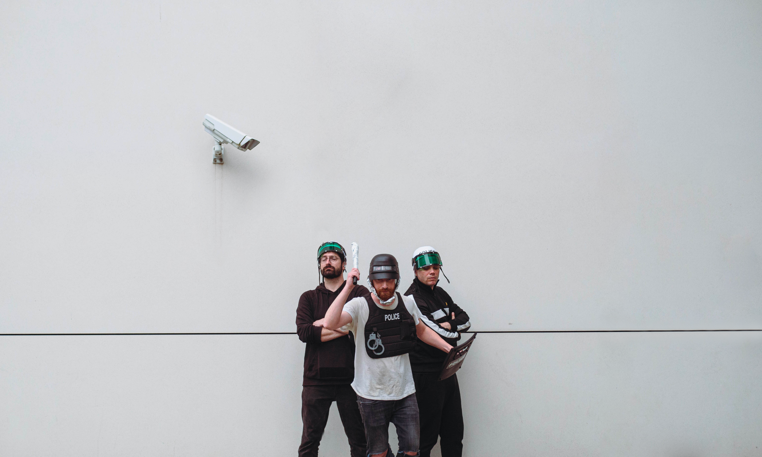 Trois personnes en tenue de police anti-émeute avec casques et boucliers, devant un mur blanc, avec une caméra de surveillance.