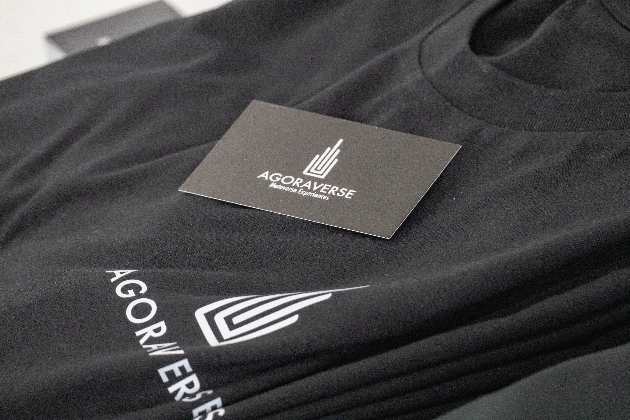 Un t-shirt noir avec le logo et le nom 'AGORA VERSE Métaverse Experiences' imprimés en blanc, avec une étiquette noire à logo posé dessus.