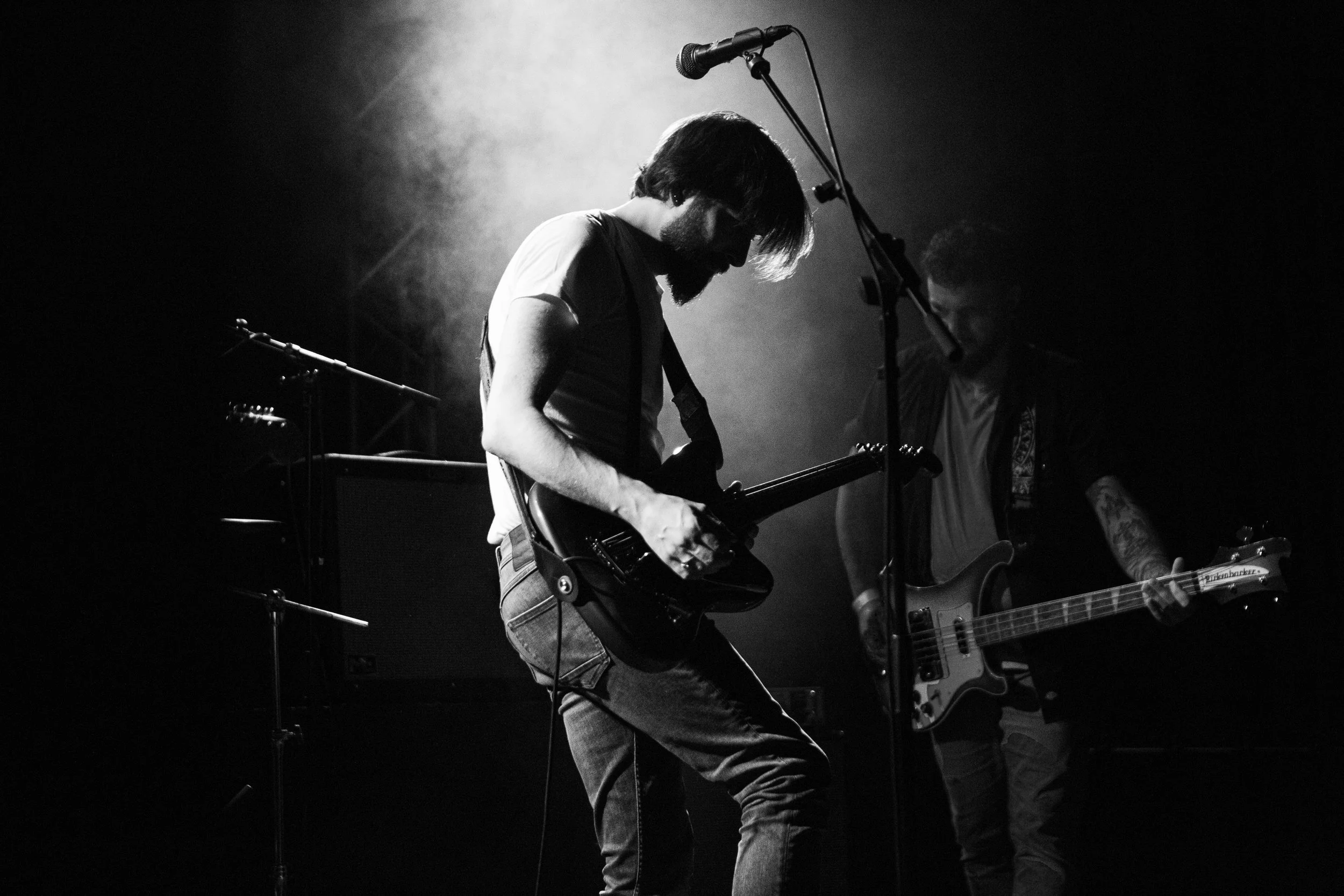 Deux musiciens jouent de la guitare électrique sur scène, en noir et blanc, avec un éclairage sombre et fumée derrière.