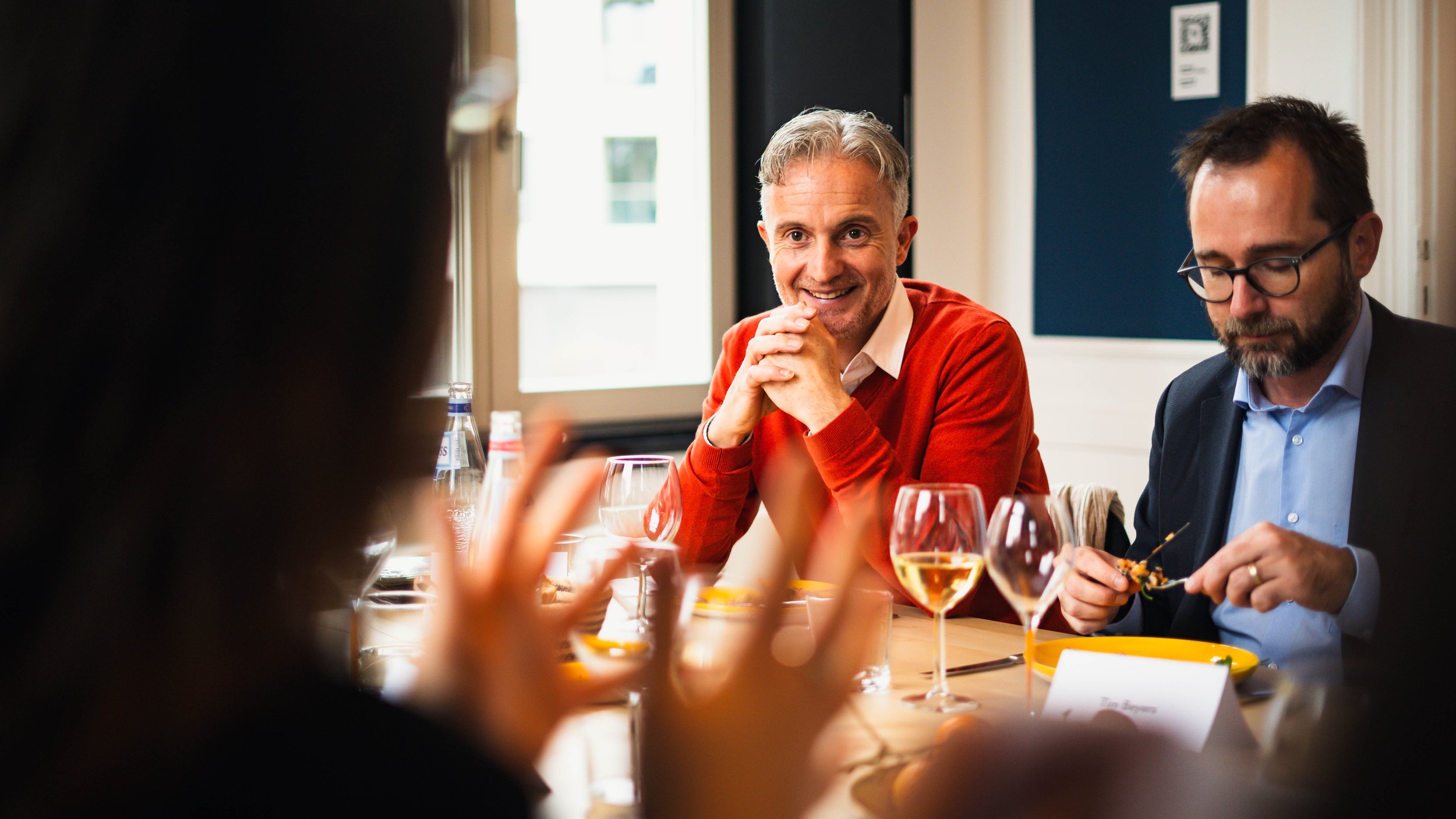 Groupe de personnes en réunion autour d'une table, souriant et conversant, avec des verres de vin et de l'eau, dans une salle bien éclairée par une fenêtre.