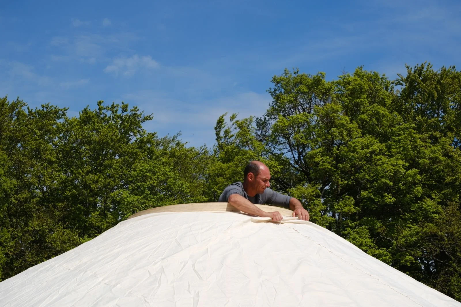 Un homme s'affaire à tisser ou à préparer une grande pièce de tissu blanc en plein air, avec un arrière-plan de ciel bleu et de nombreux arbres verts. Revetement toile intérieur de yourte 