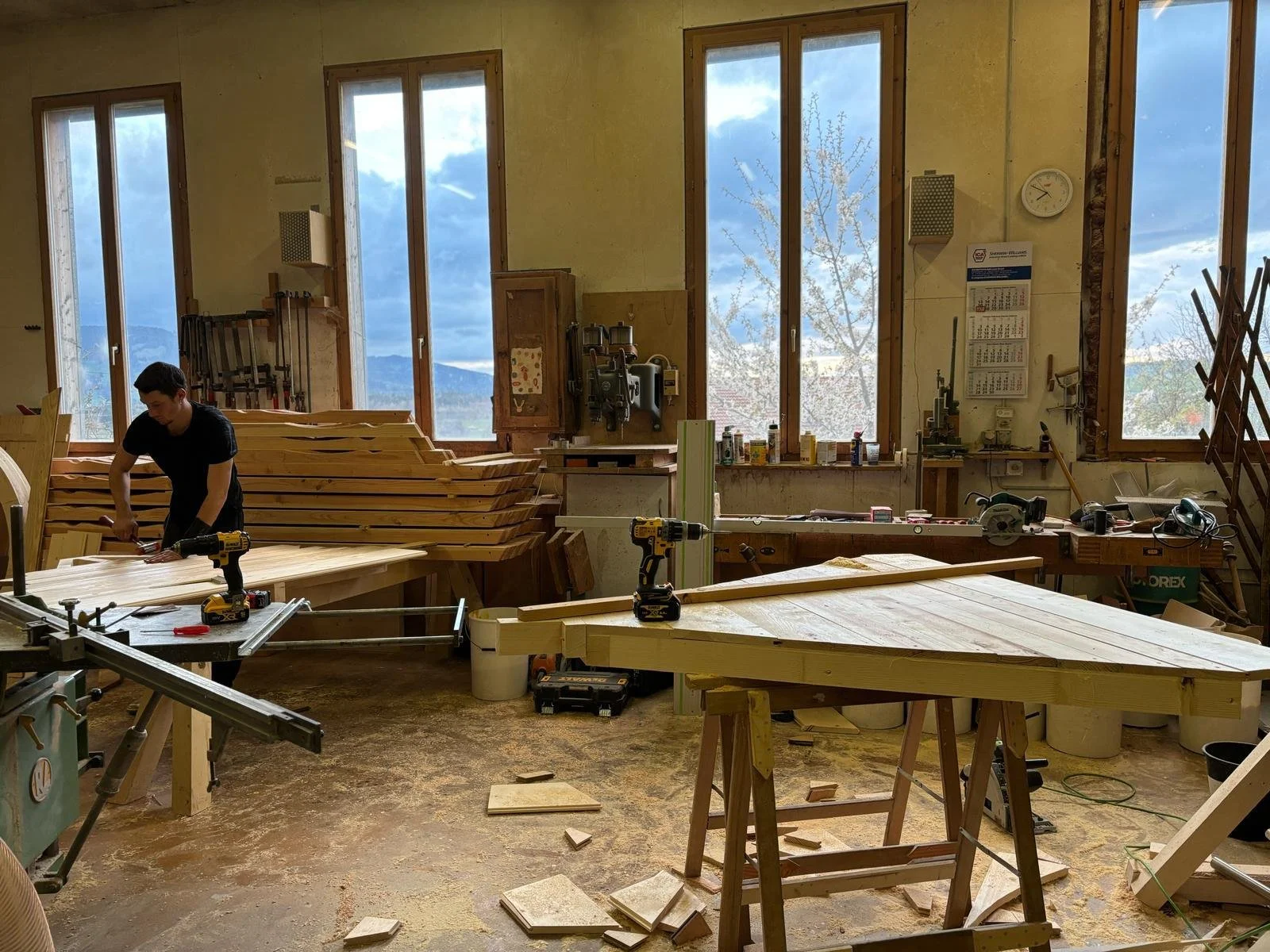 Un atelier de menuiserie avec un homme travaillant sur du bois, entouré de plans de travail, d'outils et de matériel de menuiserie, avec des fenêtres laissant entrer de la lumière naturelle.