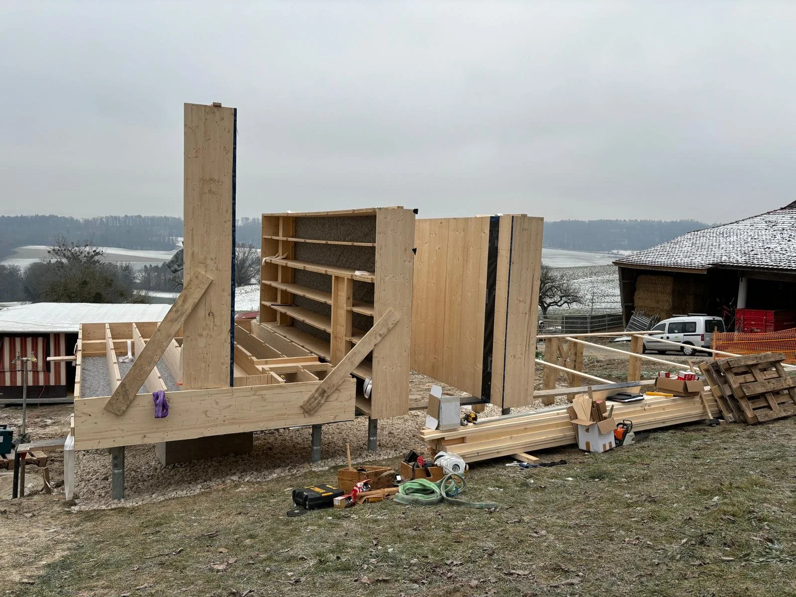 Construction en cours d'une structure en bois dans une zone rurale avec un paysage enneigé en arrière-plan, outils et matériaux de construction dispersés autour. Structure portante d'une habitation, sol. 