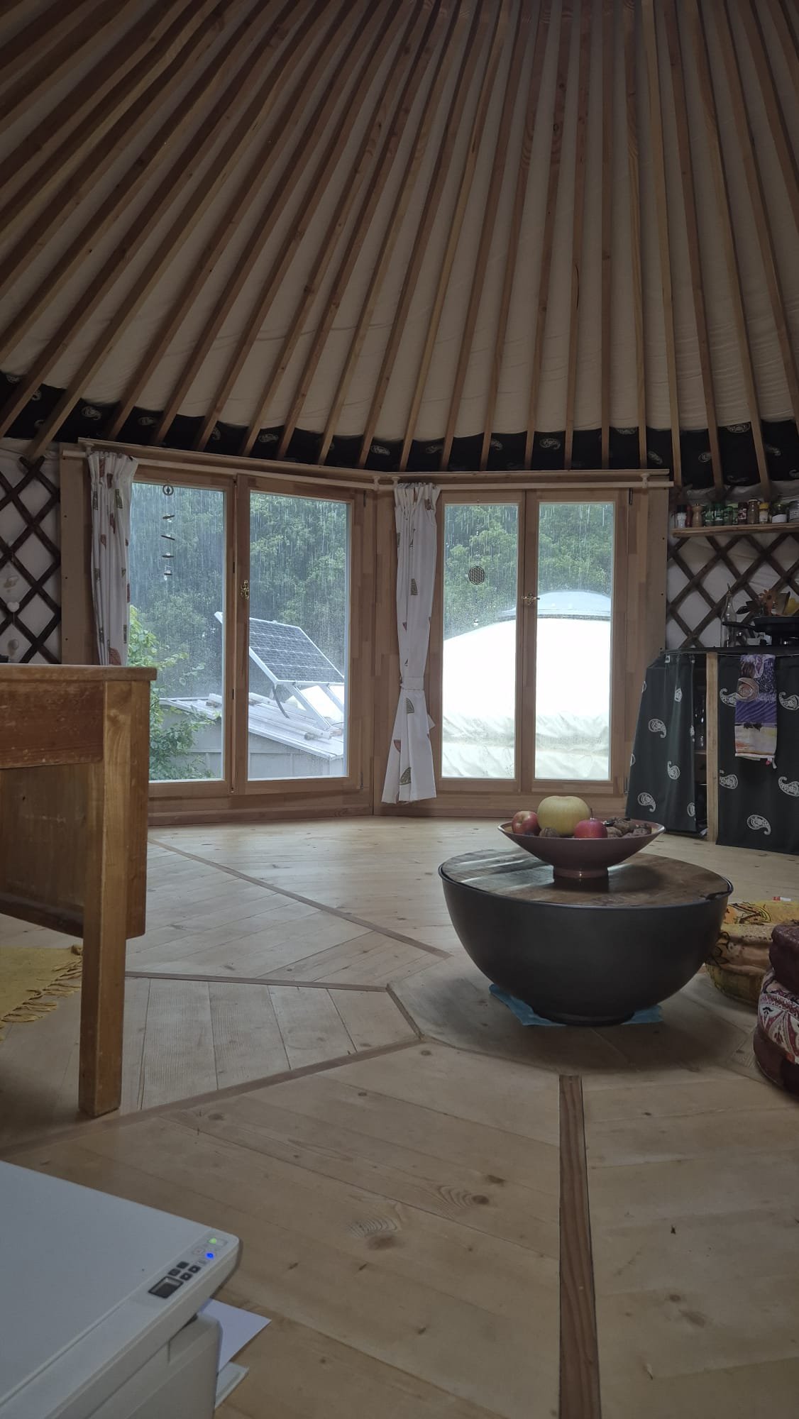 Intérieur d'une yourte en bois, avec vue sur l'extérieur à travers une grande fenêtre, décorée avec des rideaux blancs, une table basse avec un bol de fruits, et une étagère avec des bocaux.