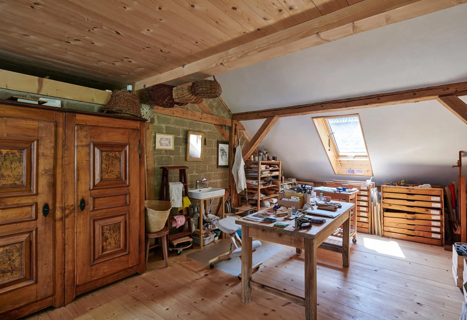 Une pièce d'atelier ou de bureau avec un plafond en bois, un mur en pierres apparentes, un bureau en bois avec des objets dispersés, une étagère en bois remplie de papiers, une fenêtre de toit laissant entrer la lumière, et des meubles en bois massif.