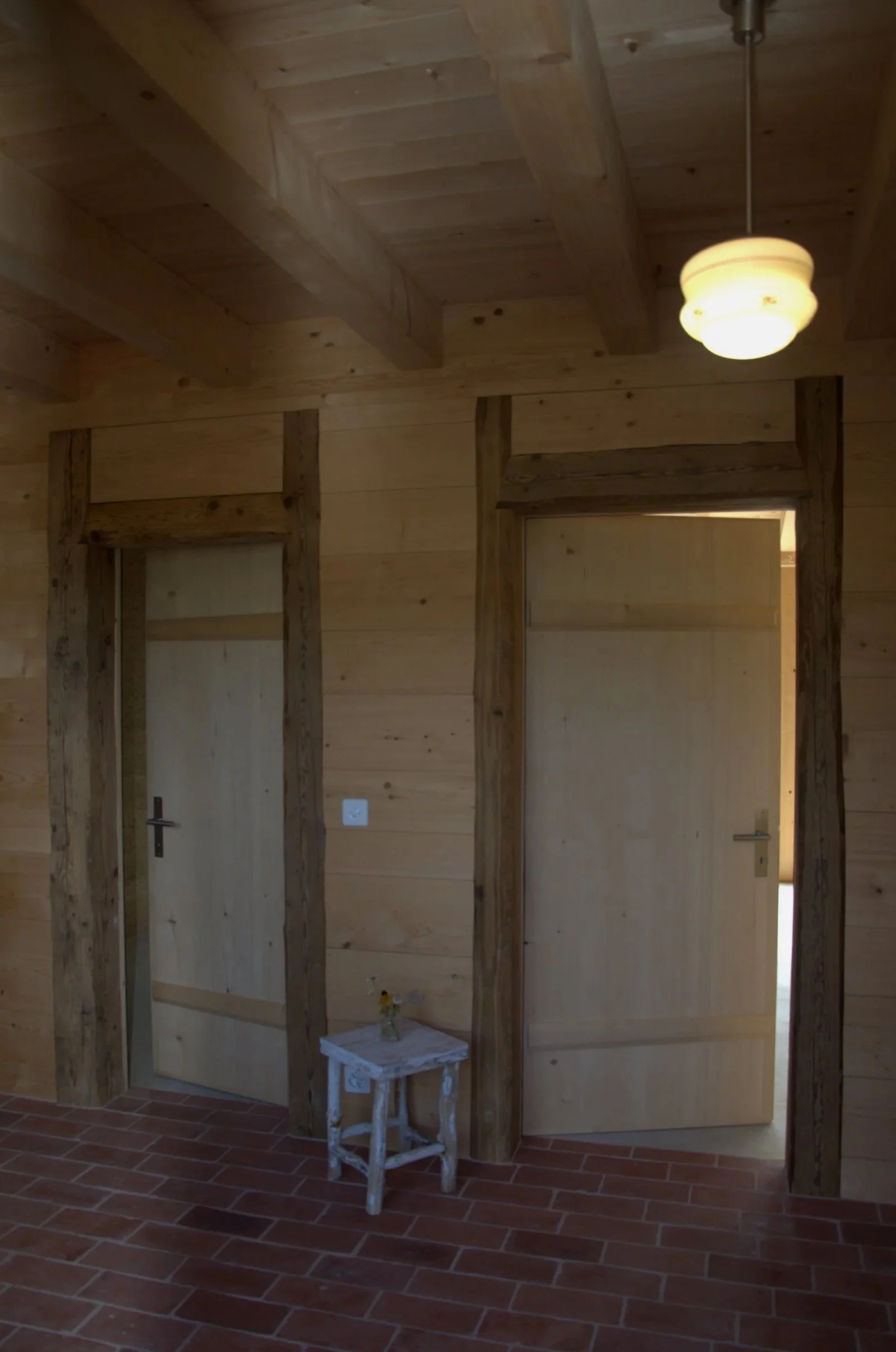 Deux portes à Eparse en bois dans une pièce avec murs en bois clair, sous un plafond en bois avec poutres apparentes et un luminaire, un petit tabouret blanc avec une petite plante est posé au sol. Transformation Habitation. 