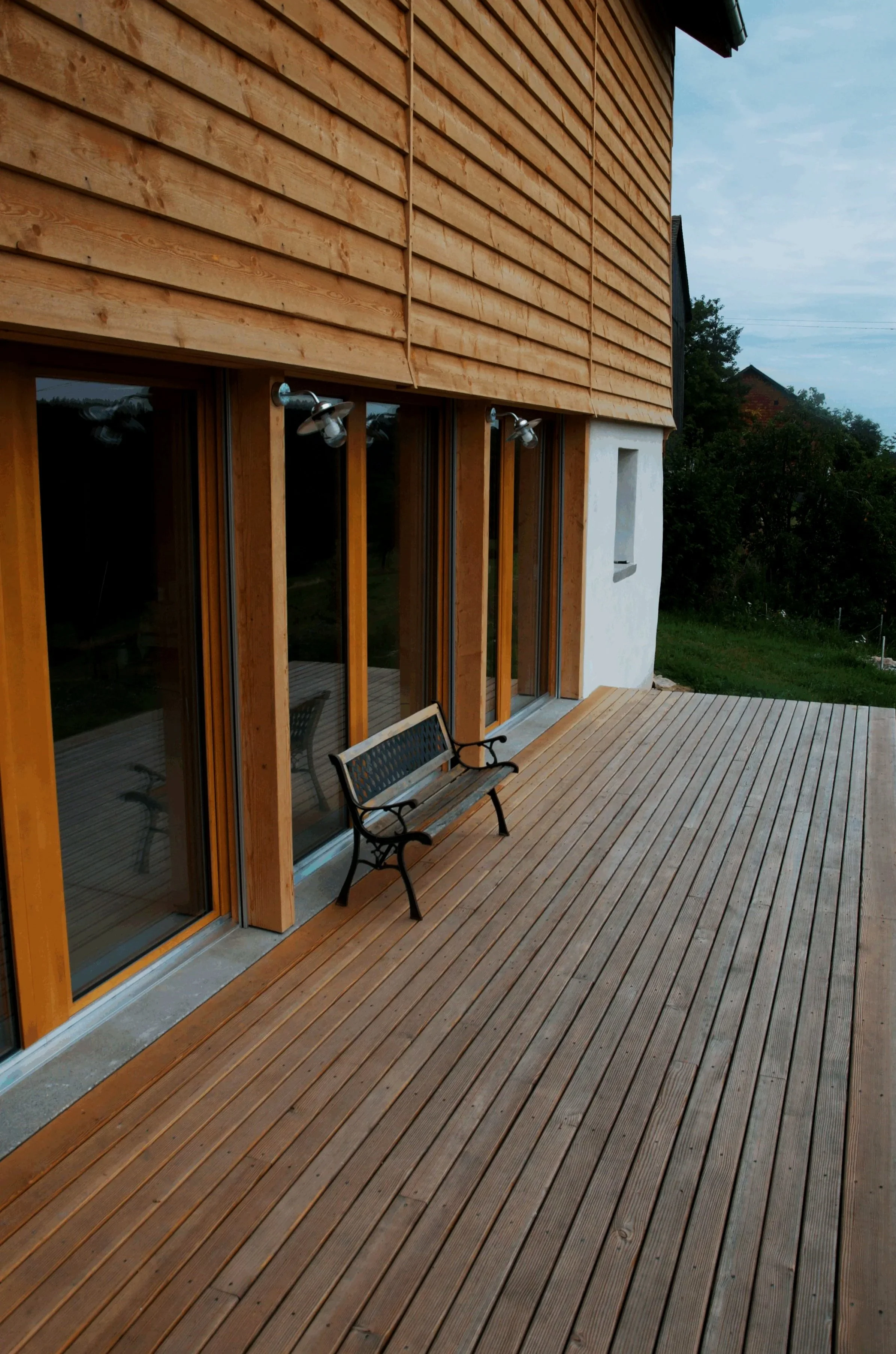 Une terrasse en bois avec un banc en métal devant une maison en bois avec de grandes fenêtres.