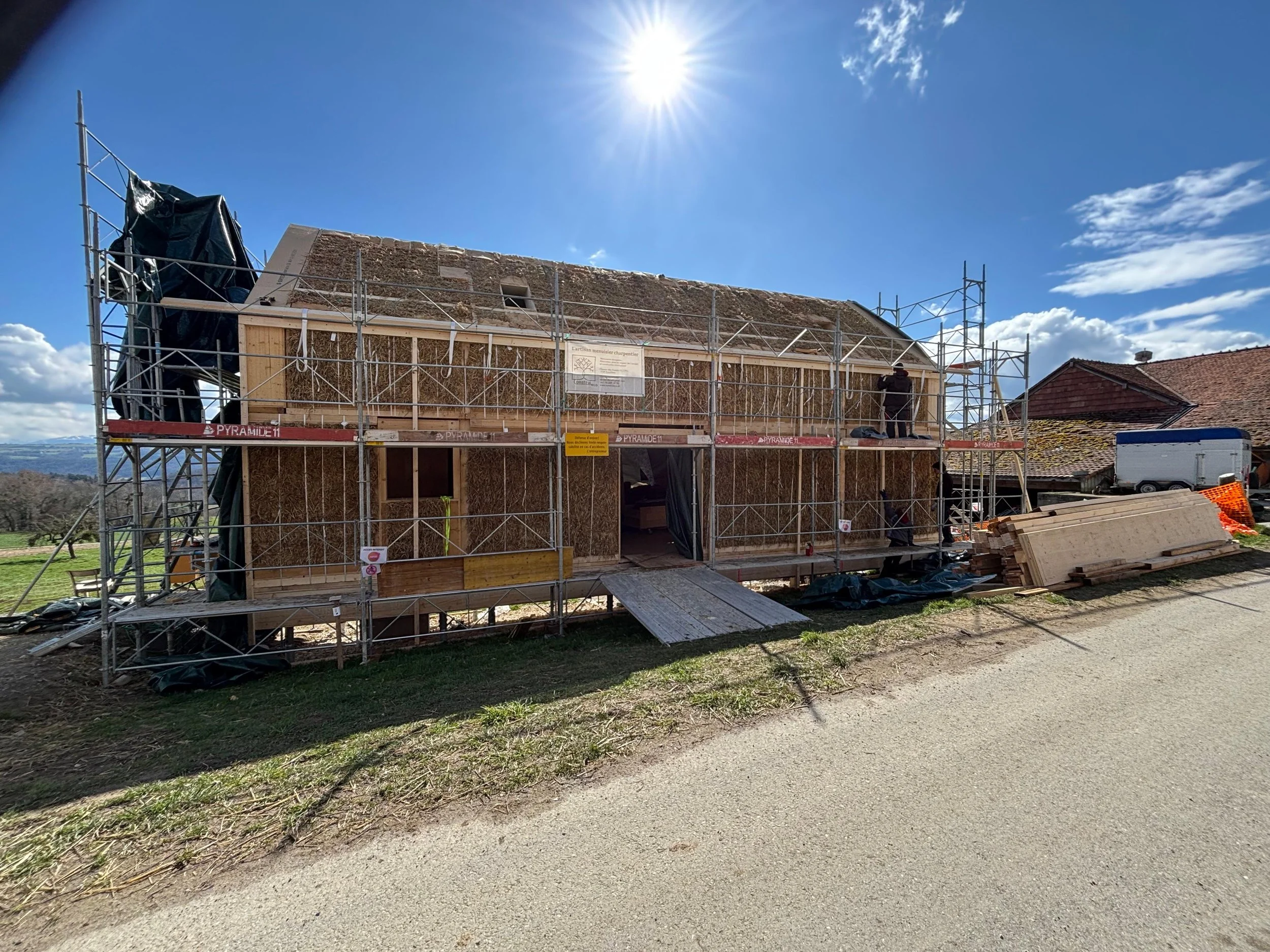 Une maison en cours de construction avec une structure en bois et des échafaudages, sous un ciel ensoleillé. Maison isolé en terra paille. ossature vb