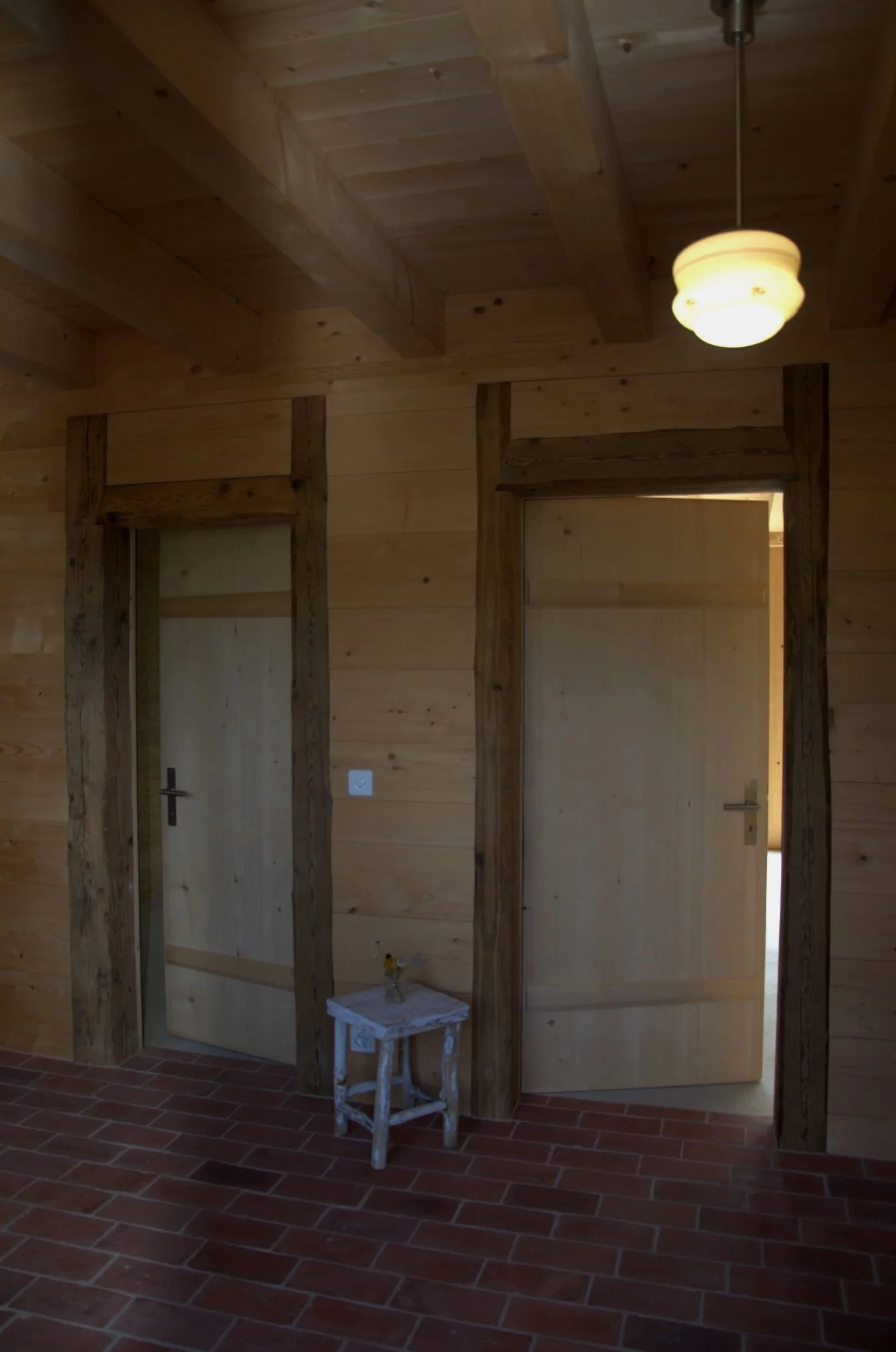 Deux portes en bois dans une pièce avec revêtement en bois, un lampadaire suspendu au plafond et un petit tabouret blanc avec une petite plante sur celui-ci.