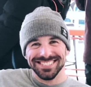 Close-up of a smiling man wearing a gray beanie and a white shirt.