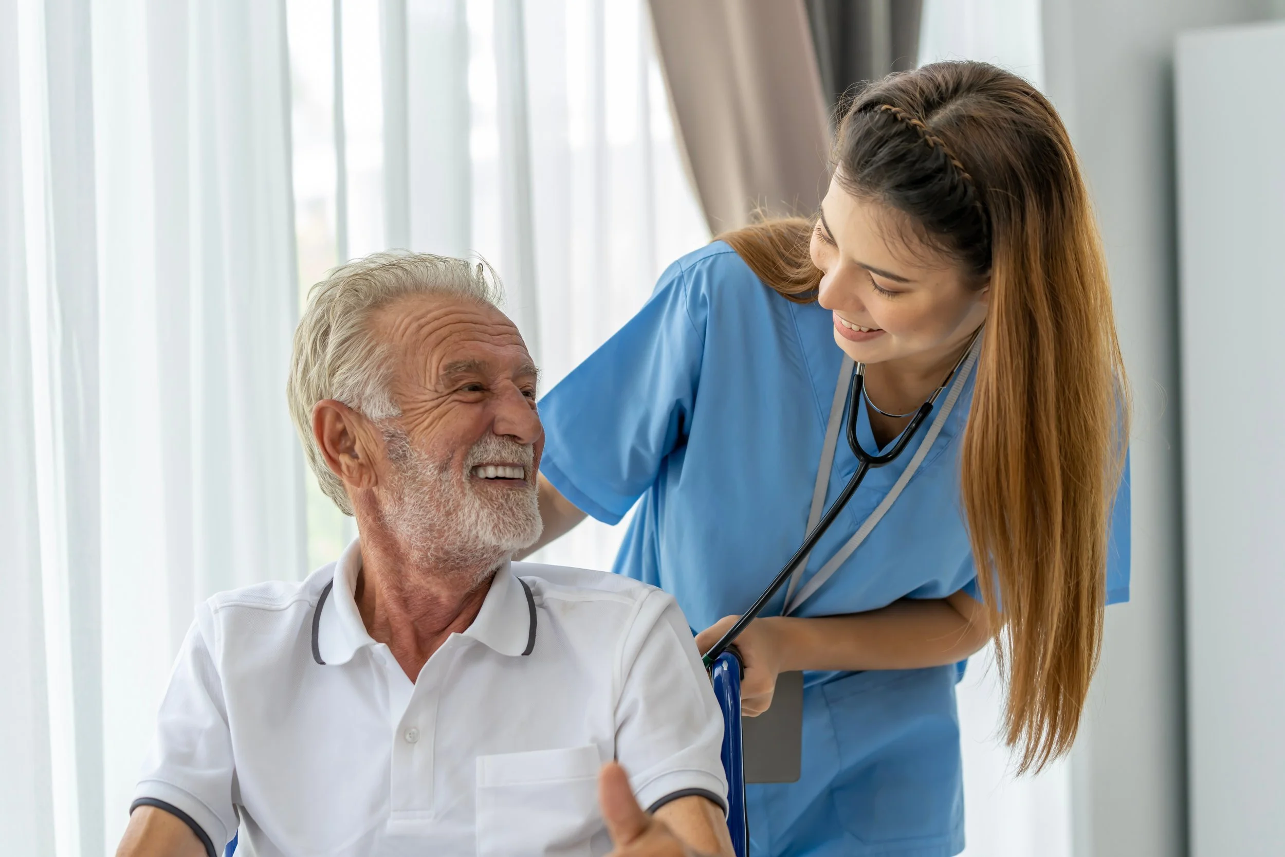 A young female healthcare professional wearing scrubs and a stethoscope smiling and talking to an older male patient who is smiling and giving a thumbs-up, in a well-lit room with large windows.