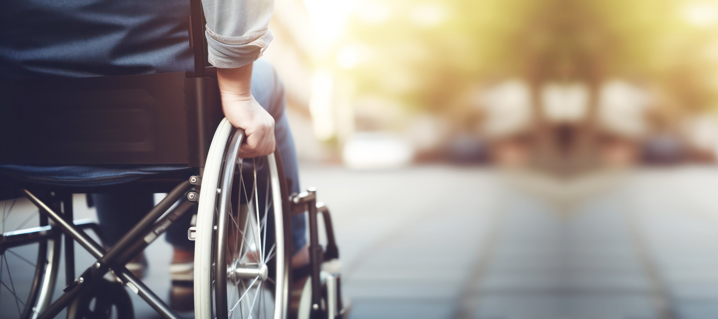 Close-up of a person in a wheelchair outdoors, holding the wheel, with sunlight and blurred background.