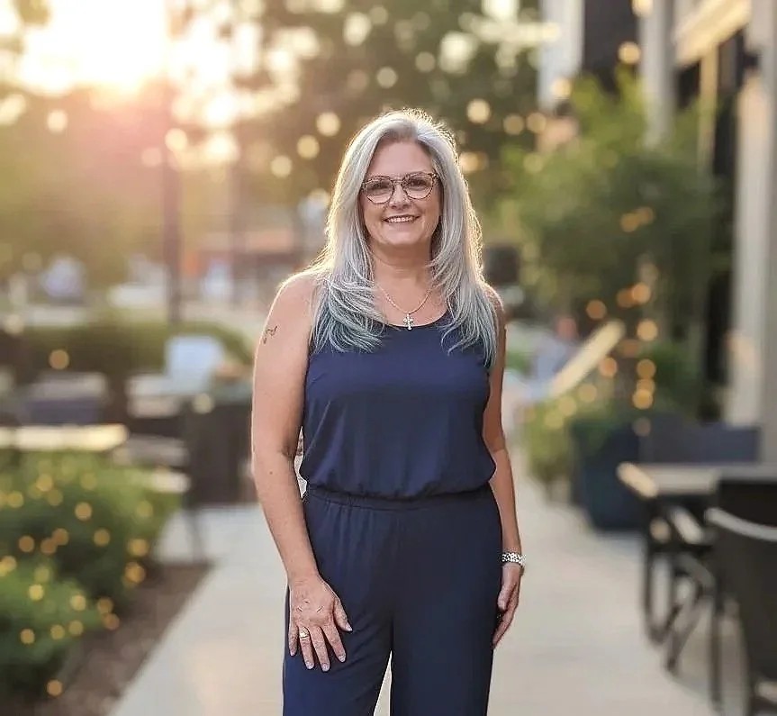 A middle-aged woman with long gray hair and glasses stands outdoors on a sidewalk, smiling, wearing a navy blue sleeveless top and matching pants, with a cityscape and trees in the background during sunset.