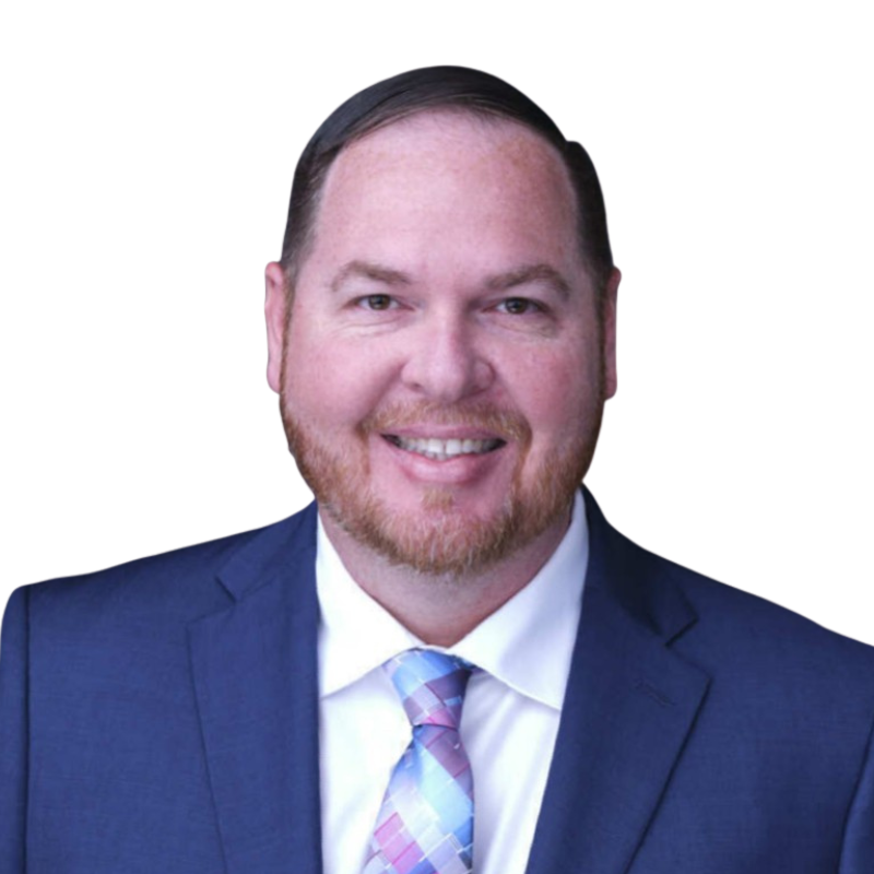 Portrait of a man with light skin, brown hair, a beard, wearing a dark blue suit, light blue dress shirt, and a checked pink and blue tie, smiling against a white background.