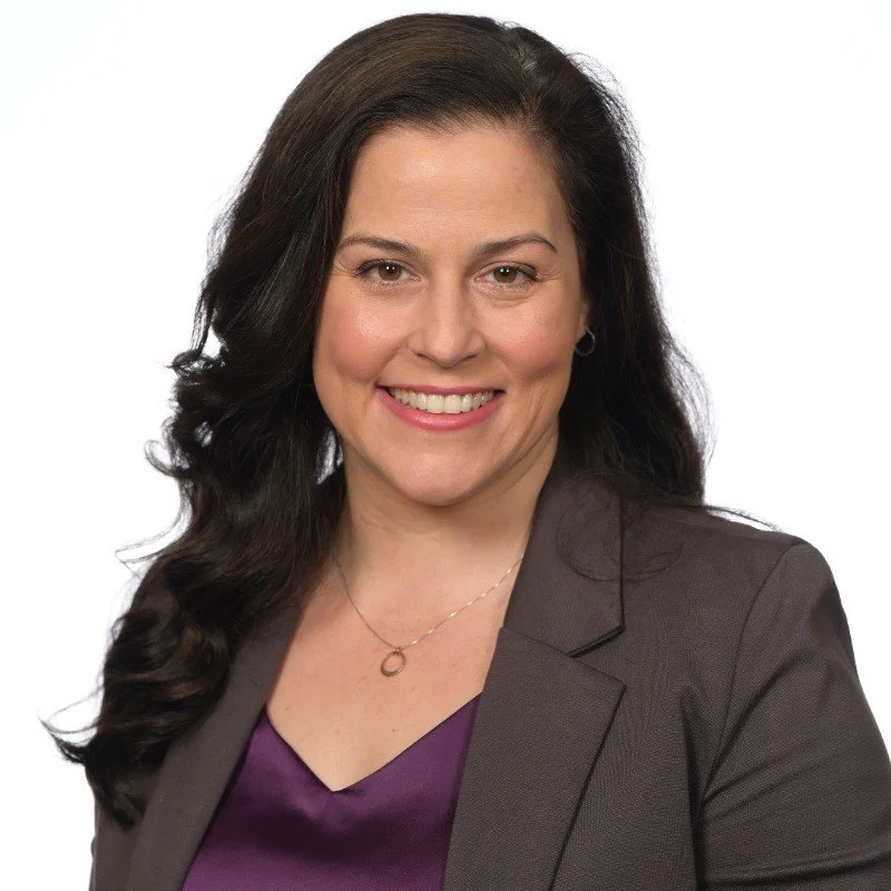 Headshot of a woman with long dark hair, smiling, wearing a dark blazer over a purple top, against a white background.