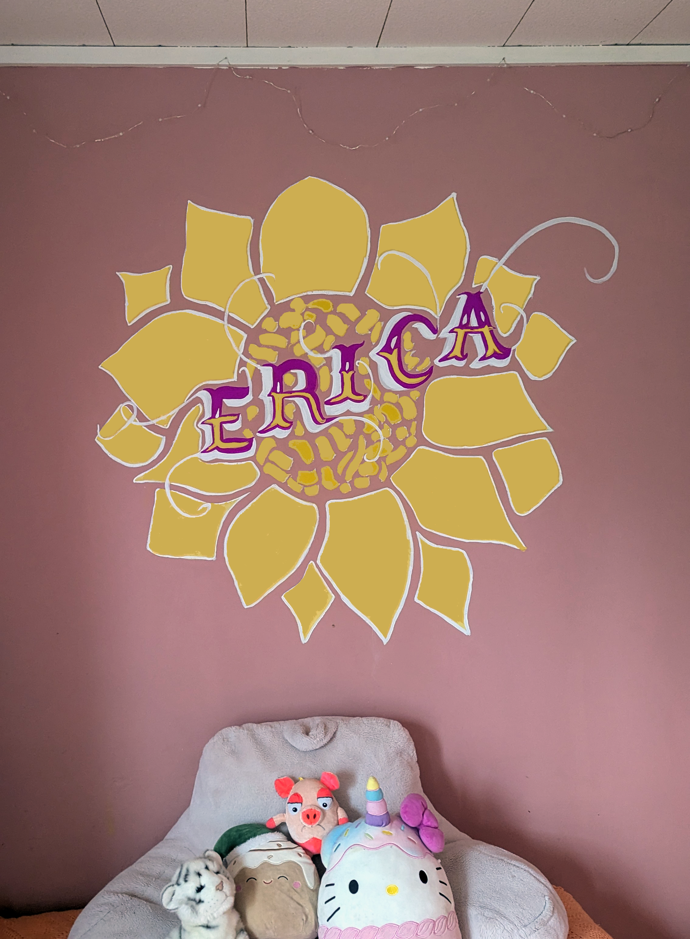 Decorative wall art featuring a large sunflower with yellow petals and a central pattern, with the name 'ERICA' written across it in purple letters. There are string lights hanging above the sunflower. Below, there is a bed or couch with plush toys including a Hello Kitty pillow, a pig plush, and other stuffed animals.