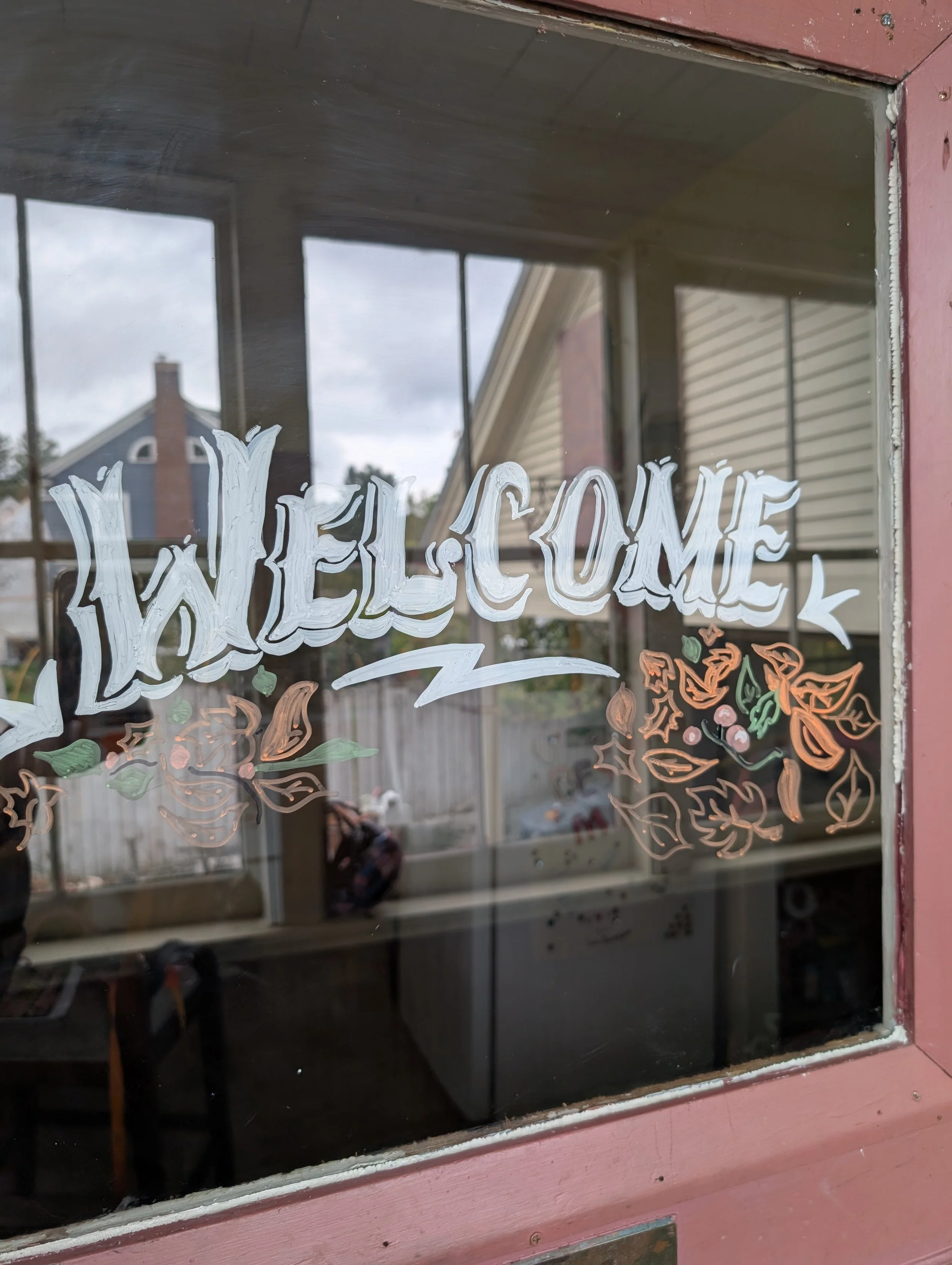 A decorative sign on a glass window saying 'WELCOME' with floral and leaf designs in orange, green, and pink colors, and a pink-painted wooden frame surrounding the window.